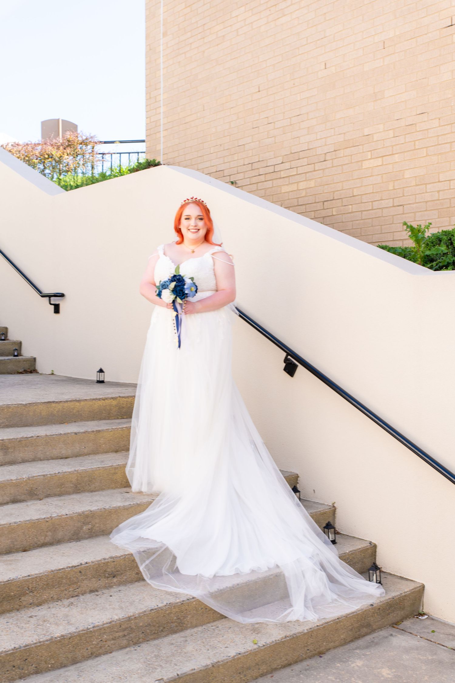 Red-haired bride in white gown holding blue bouquet poses on outdoor staircase beside brick building.