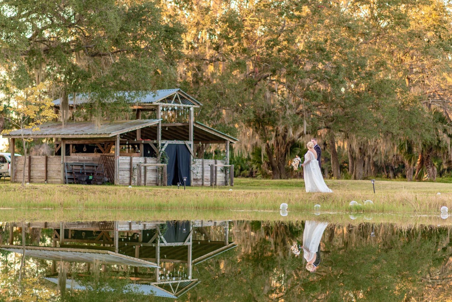Bride walks near a rustic barn reflected in a calm pond at a golden hour outdoor wedding venue.