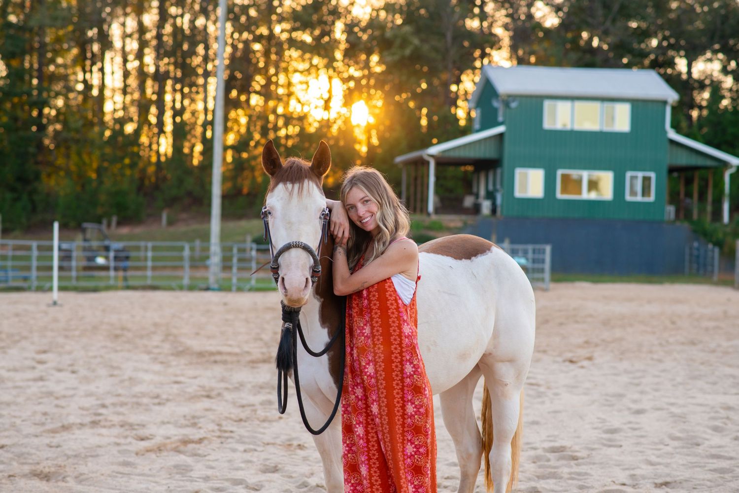 Young woman in red floral dress stands with white horse at sunset near barn.