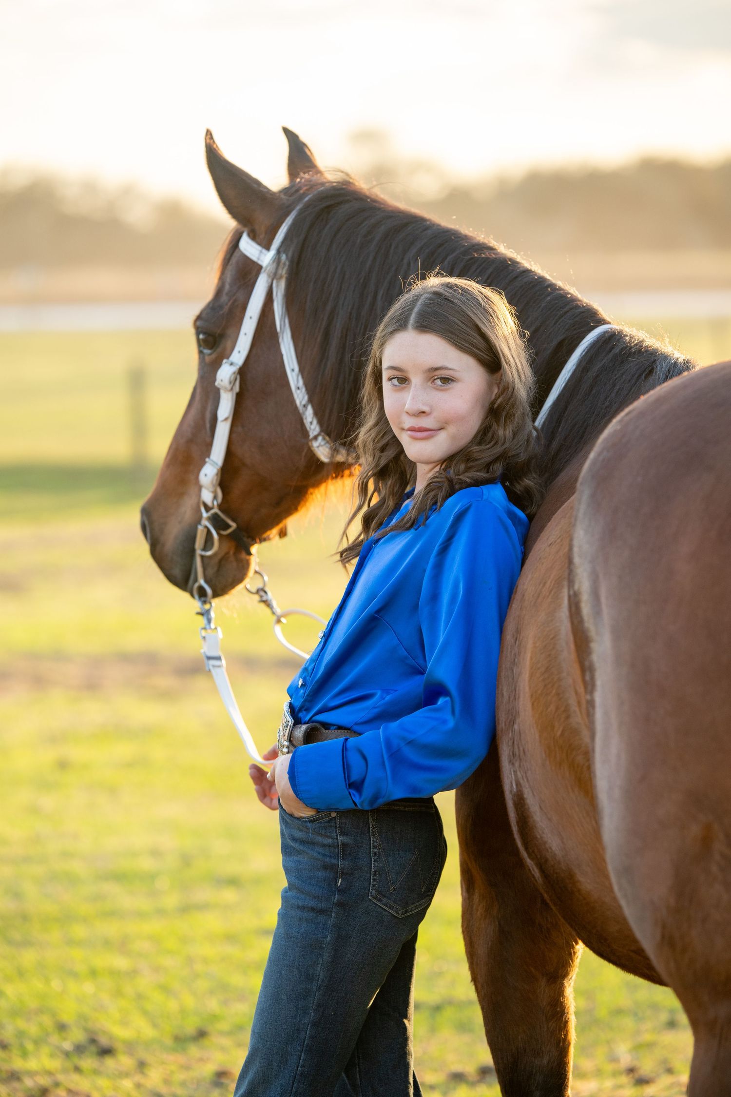 Young girl in blue jacket standing beside chestnut horse at golden hour on farm.