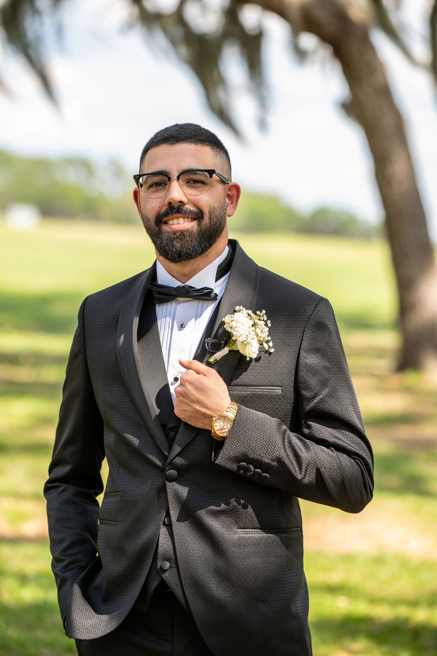 Smiling groom in a black tuxedo with boutonniere poses outdoors under a large tree.