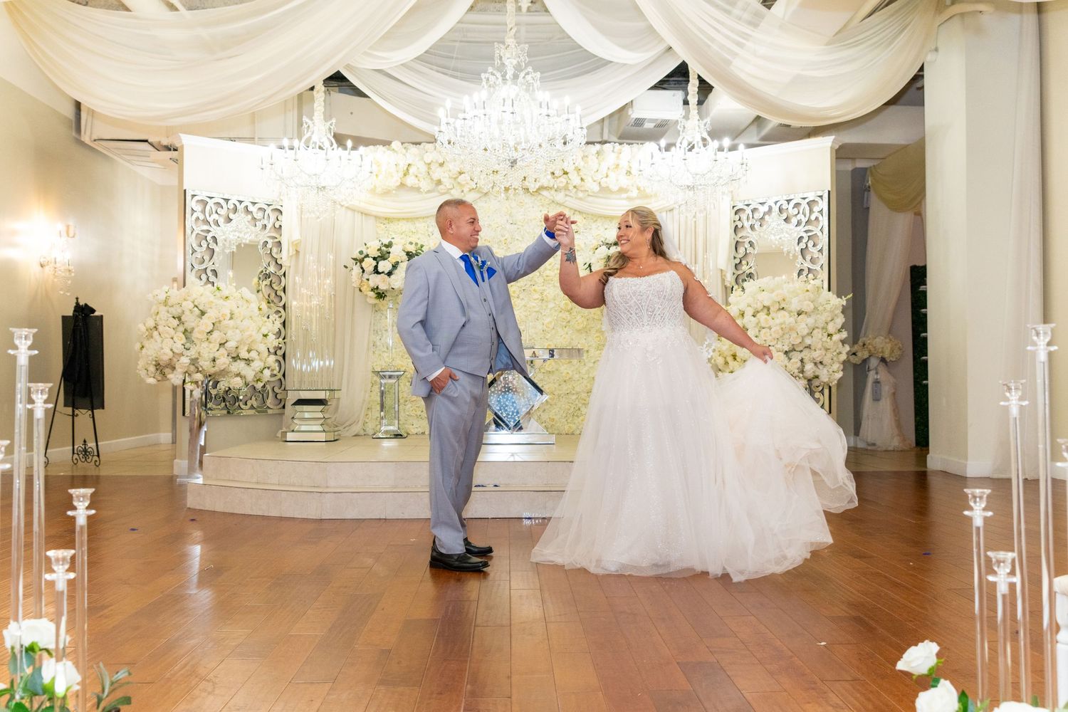 Bride and groom share first dance in elegant white wedding venue with chandelier and floral decorations.