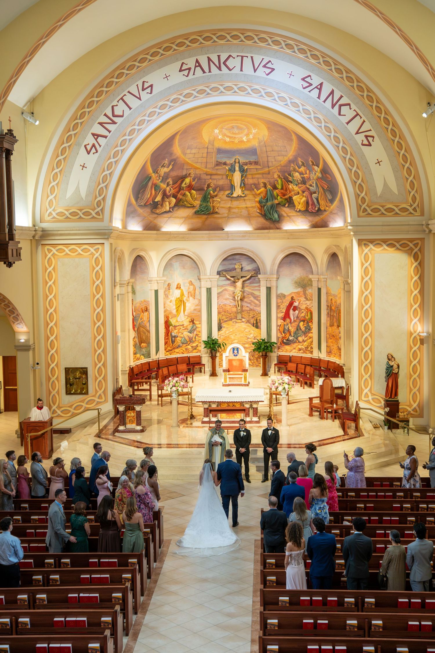 Bride walking down the aisle in a stunning Catholic church with ornate murals and Sanctus inscriptions during wedding ceremony.