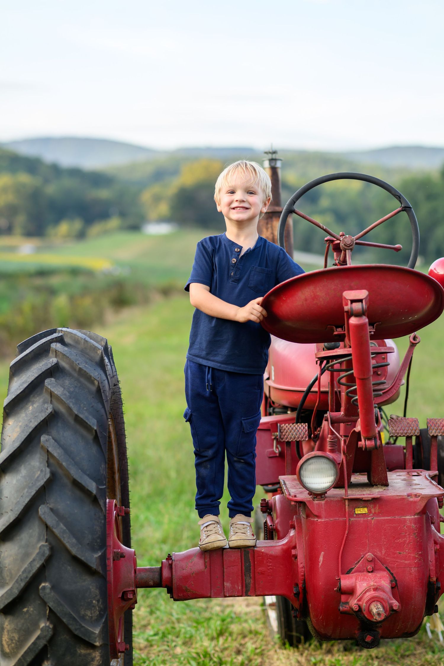 Young blonde boy in navy overalls standing on a vintage red tractor in a green farm field with rolling hills.