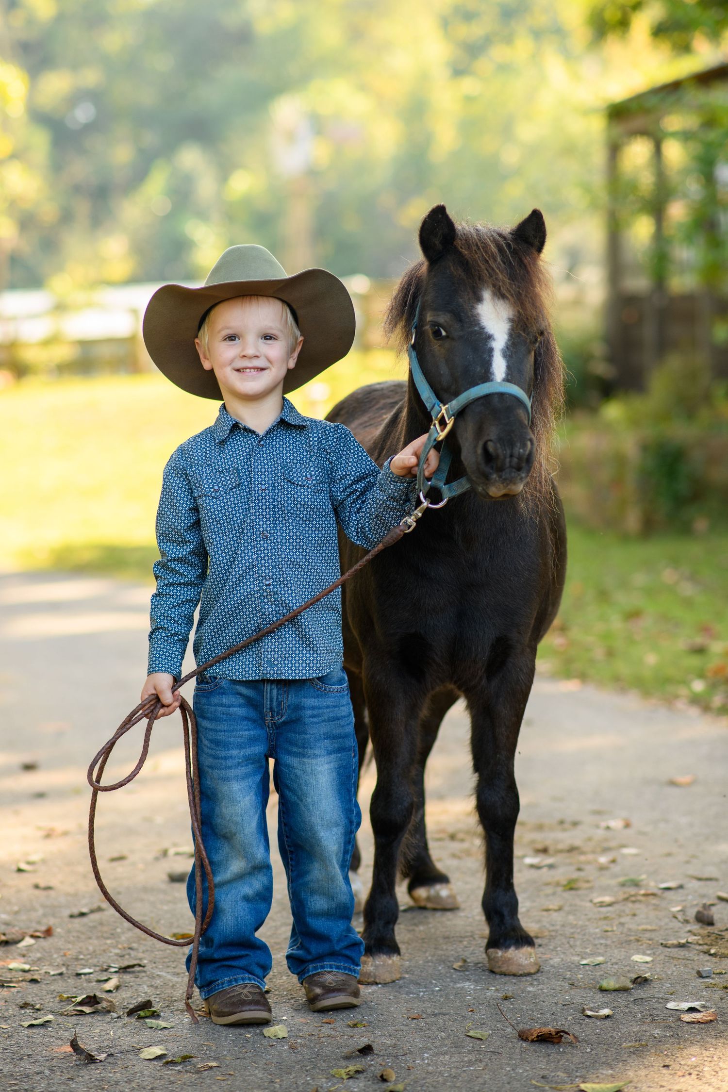 A smiling young boy in a cowboy hat and blue shirt leads a dark miniature pony on a sunny farm path.