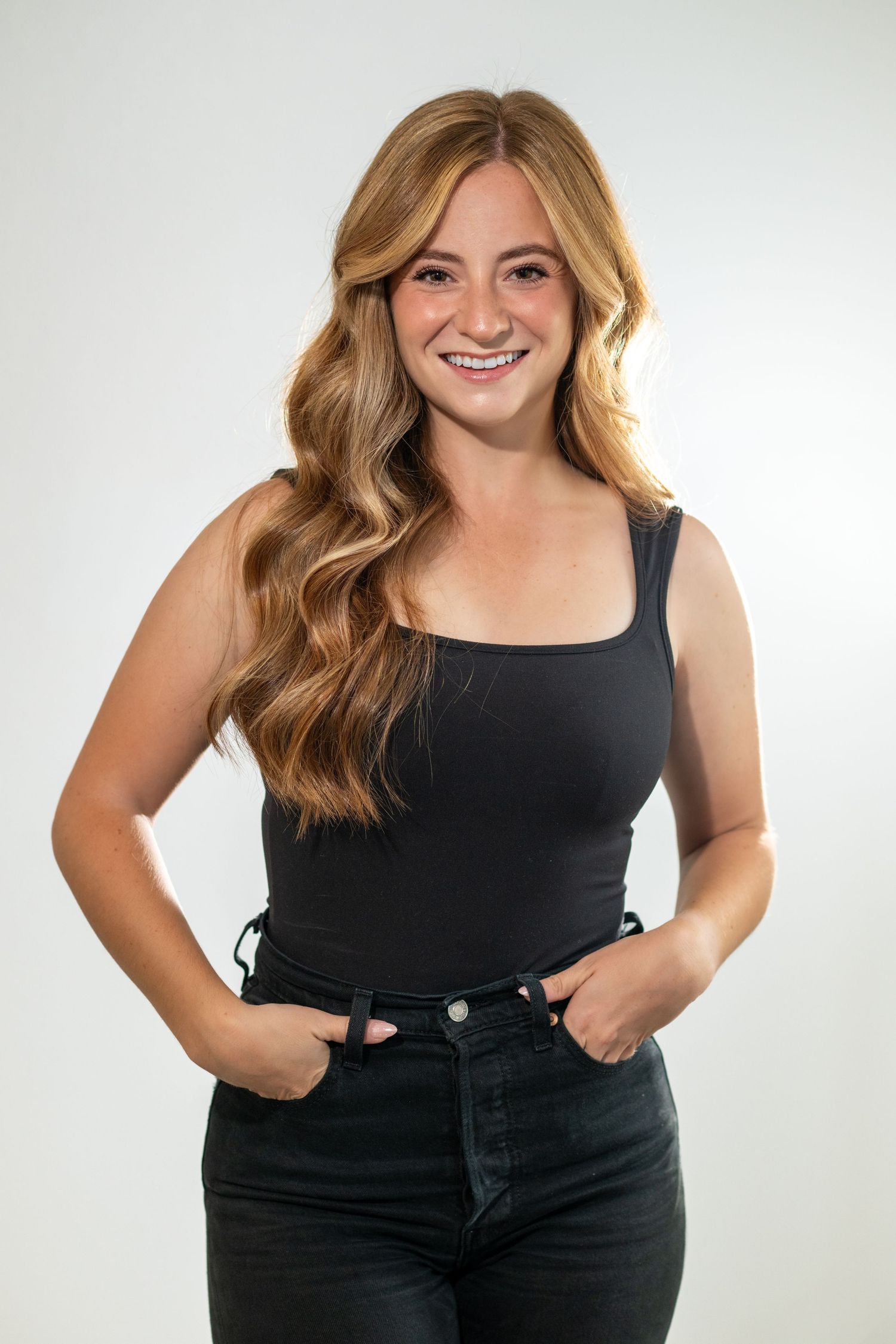 Smiling young woman with wavy blonde hair wearing a black tank top and dark jeans, posing against a white background.