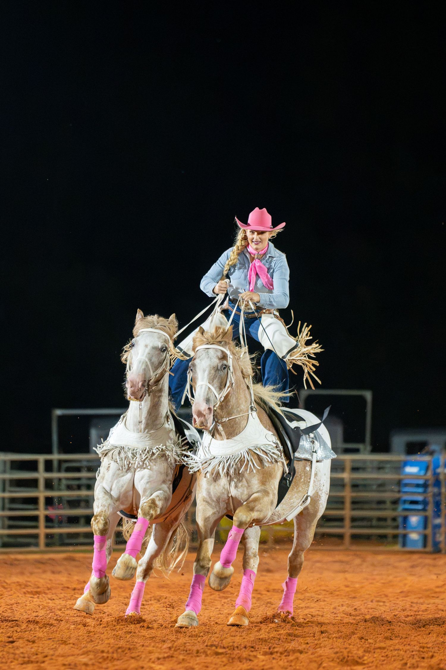 Cowgirl in pink hat rides two horses simultaneously at a nighttime rodeo event on a dirt arena.