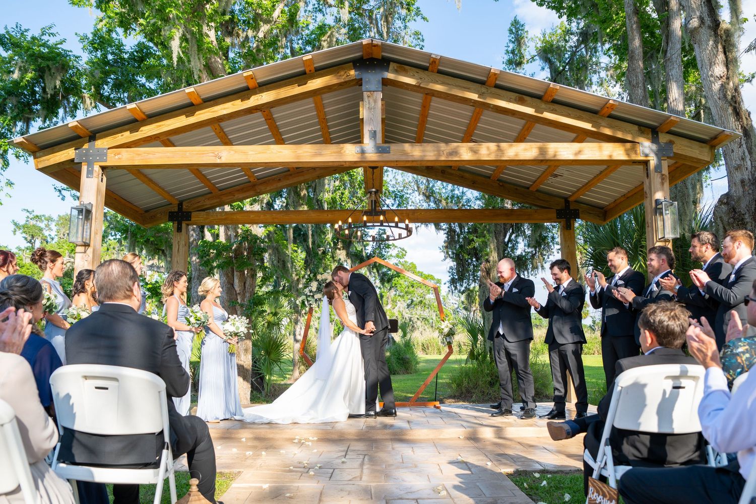 Outdoor wedding ceremony under a wooden gazebo with guests applauding the couple exchanging vows surrounded by lush greenery.