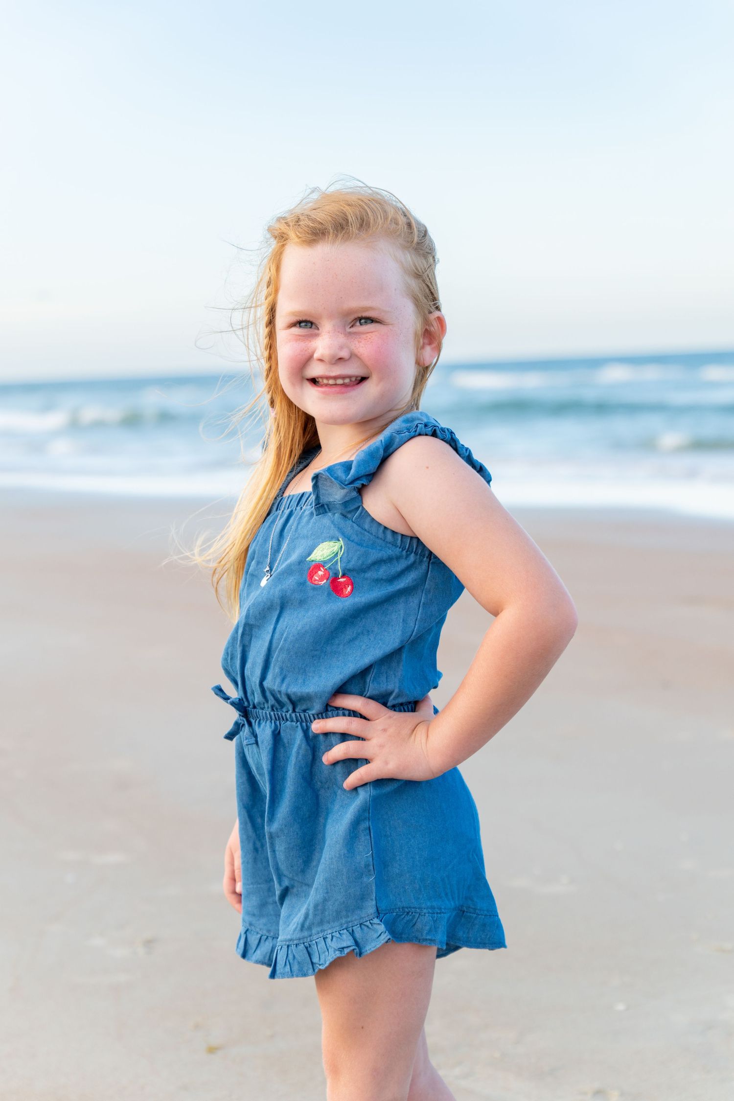 Smiling young girl in blue denim romper with cherry print poses on sandy beach with ocean waves behind her.