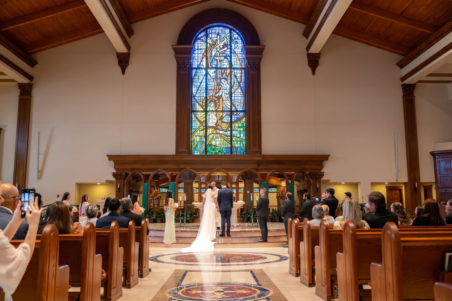 Bride and groom standing at the altar of an ornate church with a stunning stained glass window backdrop.