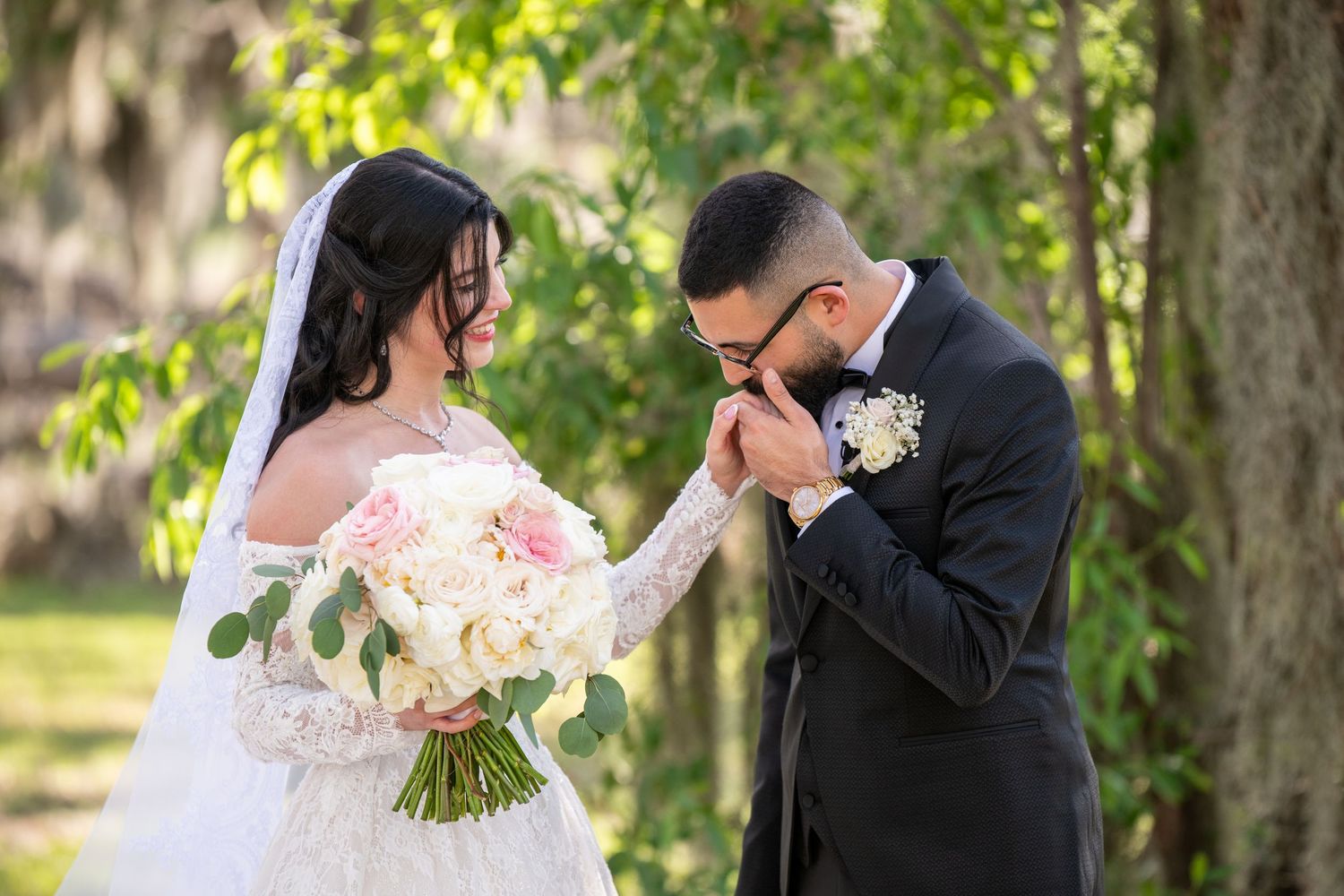 Groom gently kisses the hand of his bride holding a pink and white bouquet outdoors.