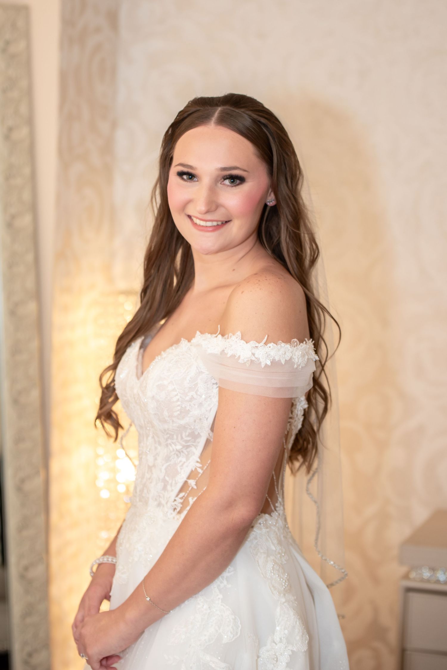 Brunette bride in off-shoulder lace ball gown smiles elegantly in softly lit bridal suite.