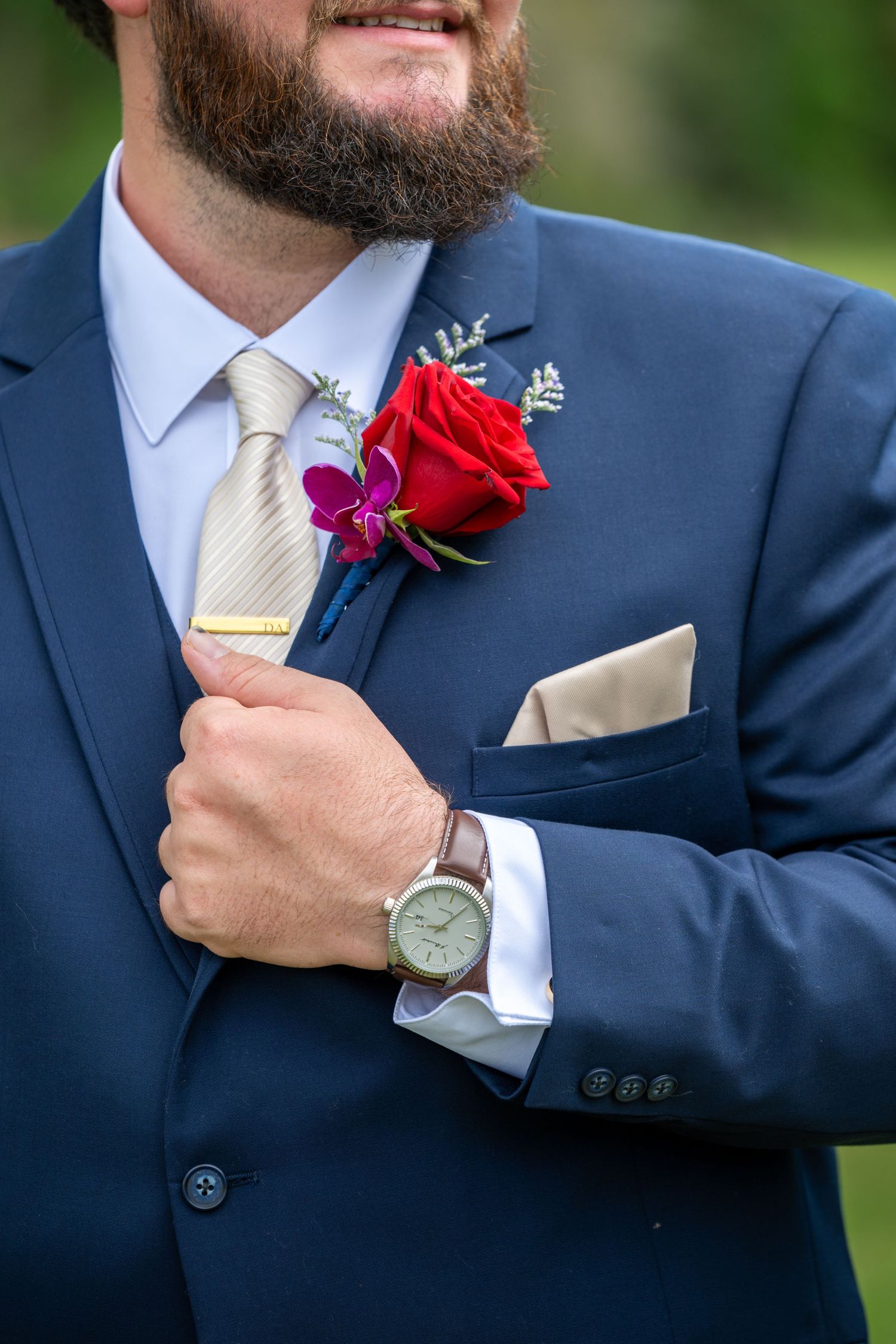 Groom in blue suit adjusting red rose boutonniere with a watch on his wrist.