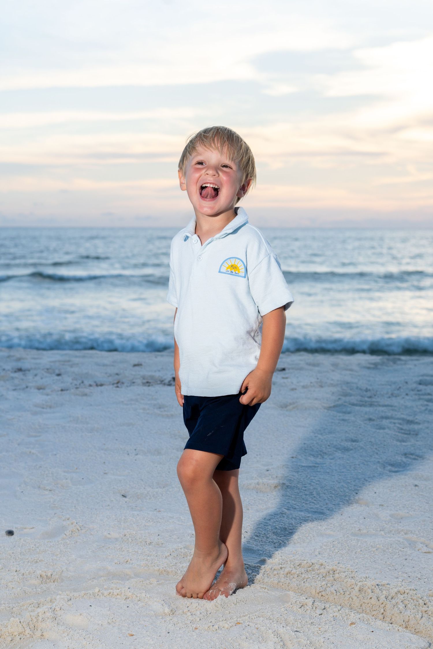 Laughing young boy in a white polo shirt standing barefoot on a sandy beach at sunset.
