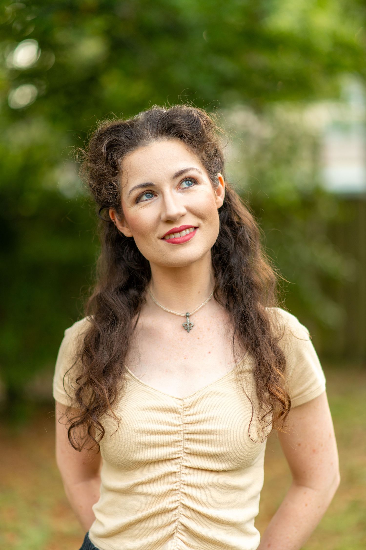 Young woman with long curly brown hair wearing a yellow top smiling outdoors with green trees behind her.