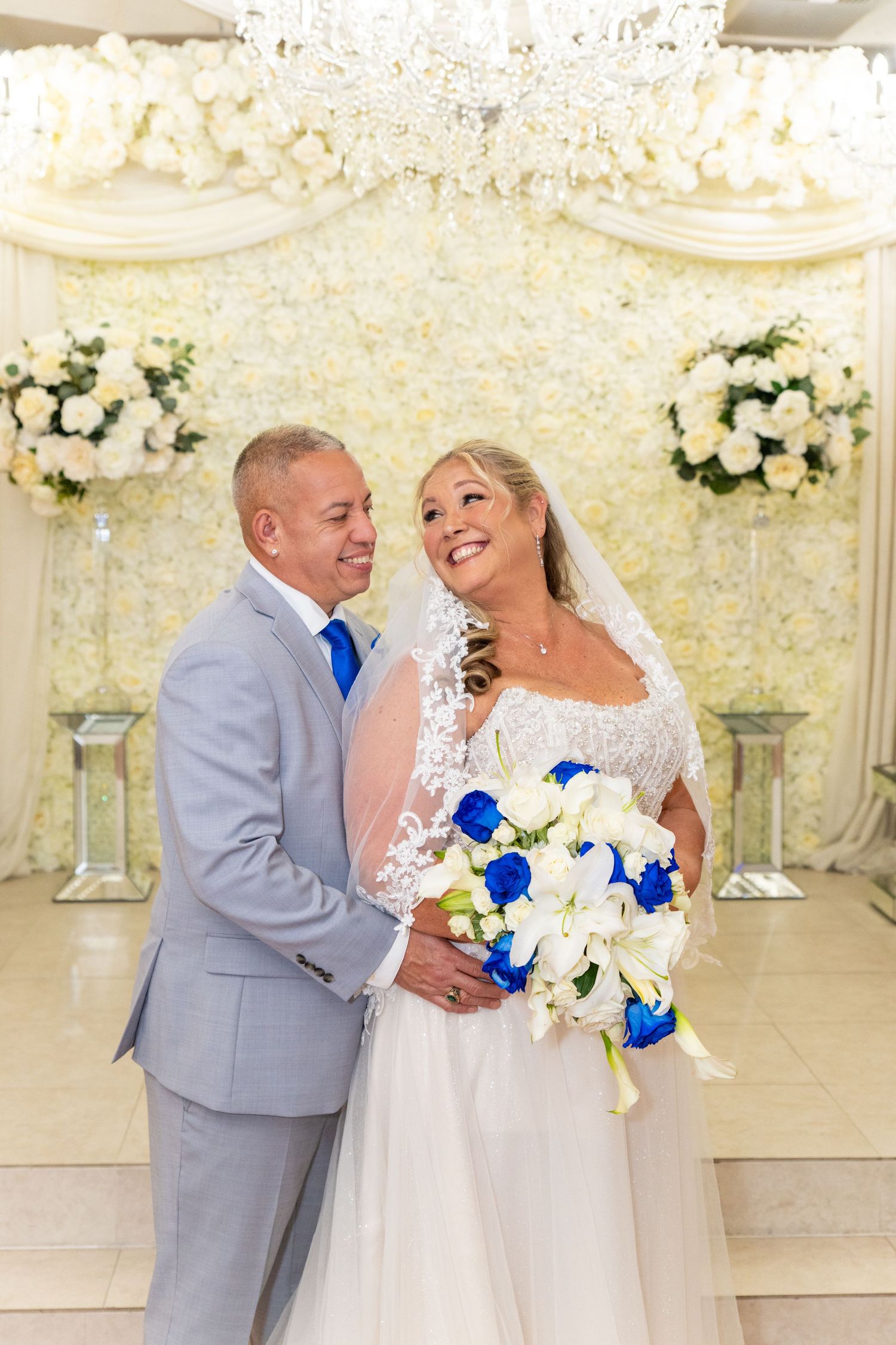 Bride and groom smile together at wedding venue with white floral backdrop, chandelier, and blue and white bouquet.