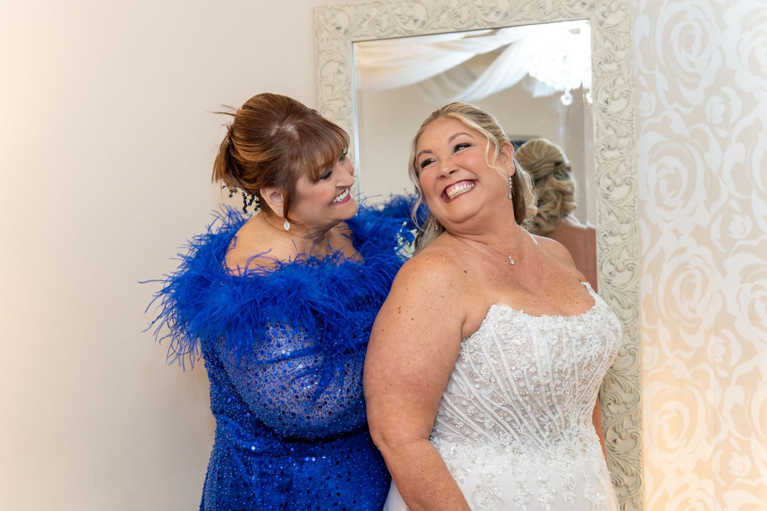 Bride in white strapless gown smiles as woman in blue feather dress helps her prepare in front of ornate mirror.