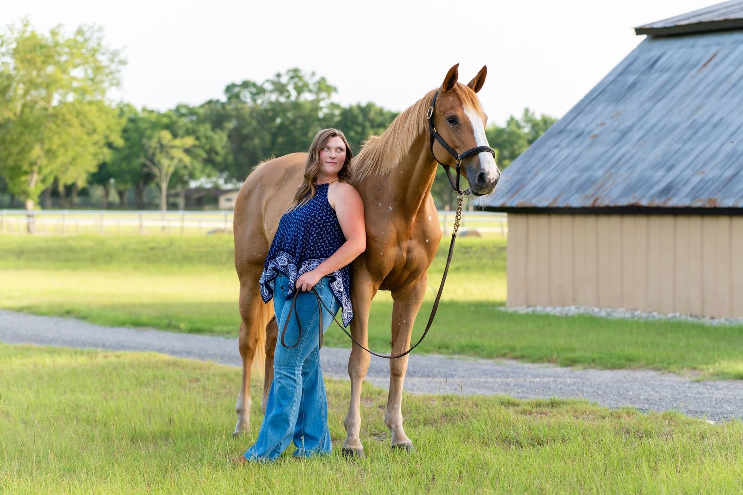 Woman in blue dress standing with a golden chestnut horse near a barn on green pasture.