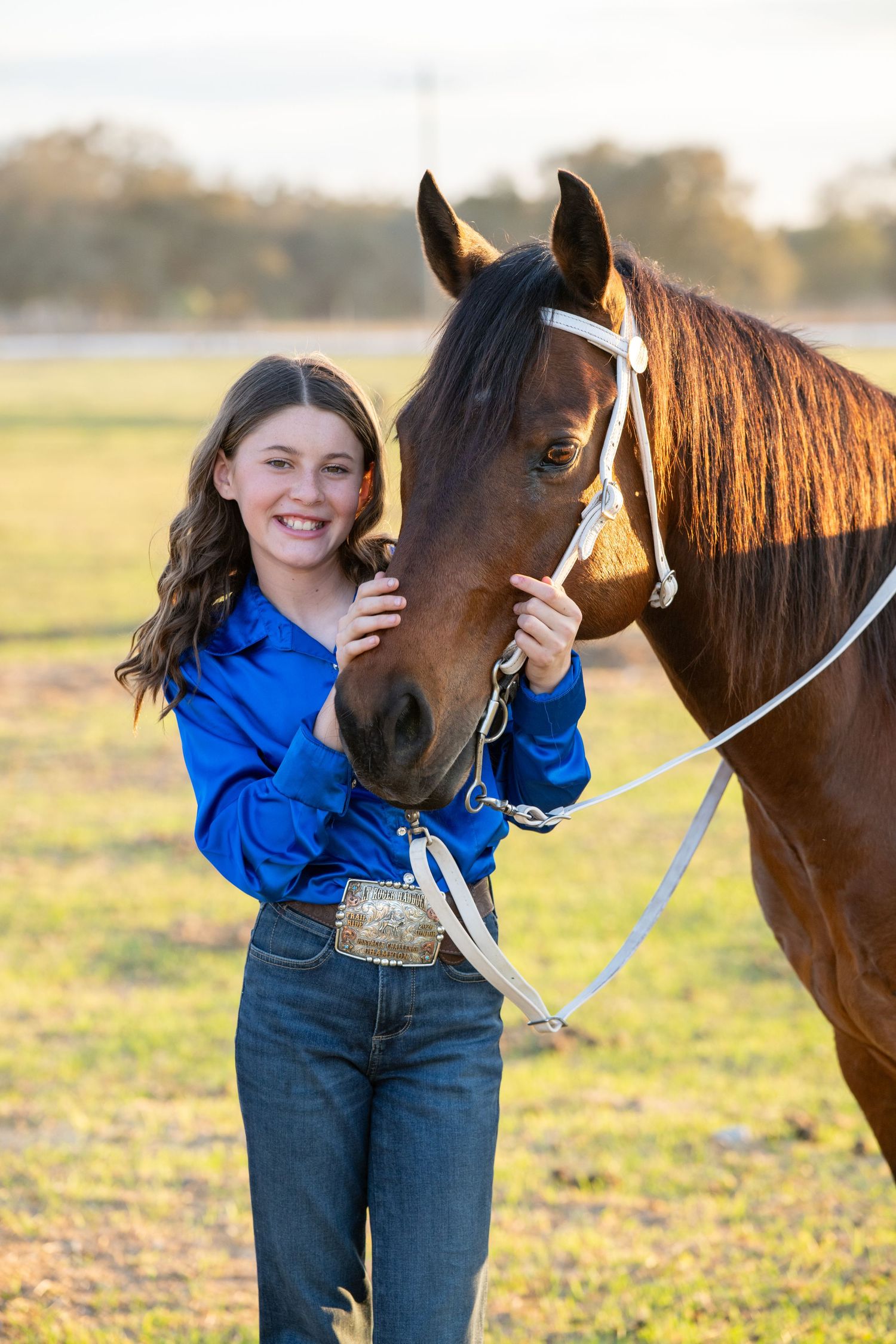 Young girl in blue western shirt smiling beside a bay horse in a golden field.