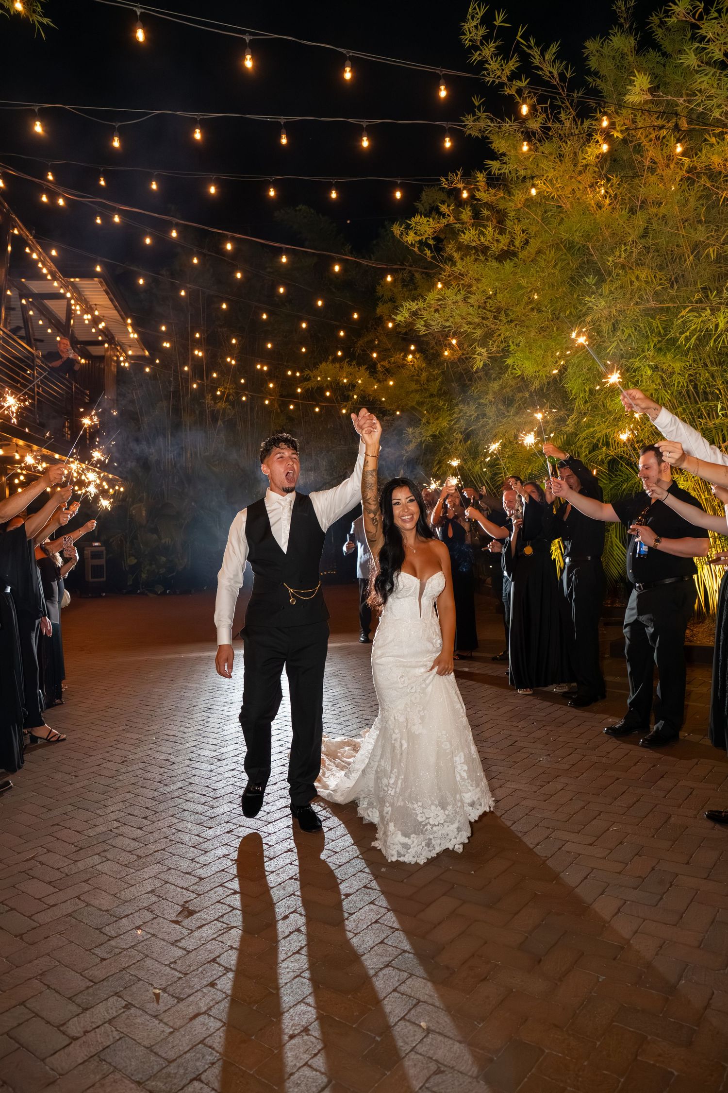 Bride and groom walk through sparkler exit under string lights at night wedding reception on brick pathway.