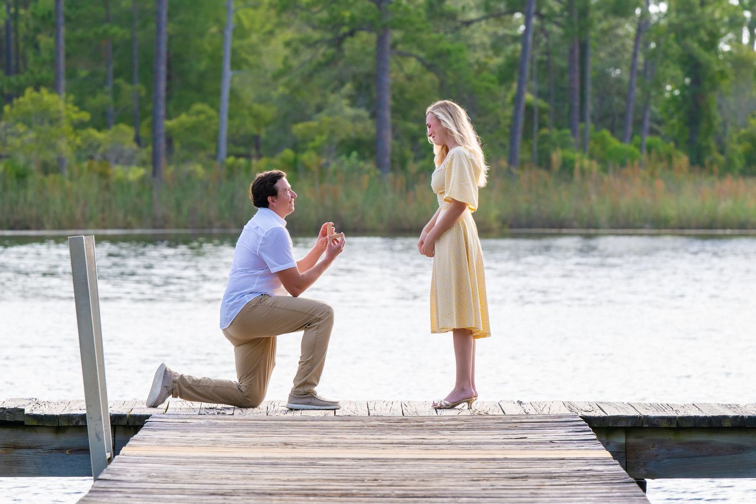 Man proposing to woman on a wooden dock by a lake, kneeling with a ring in hand on a sunny day.