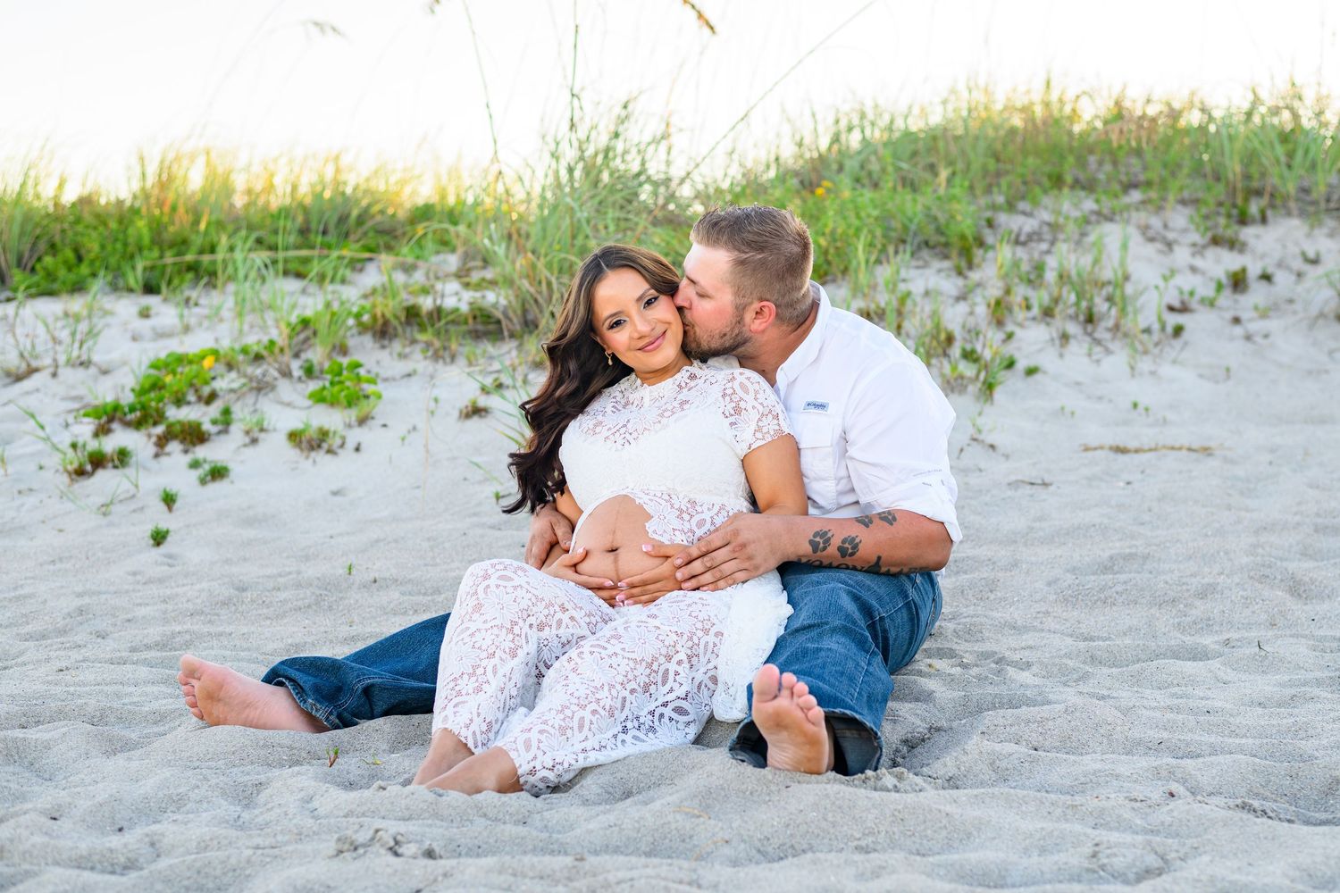 Expectant couple sits together on a sandy beach as the man kisses the woman's cheek while both rest hands on her baby bump.