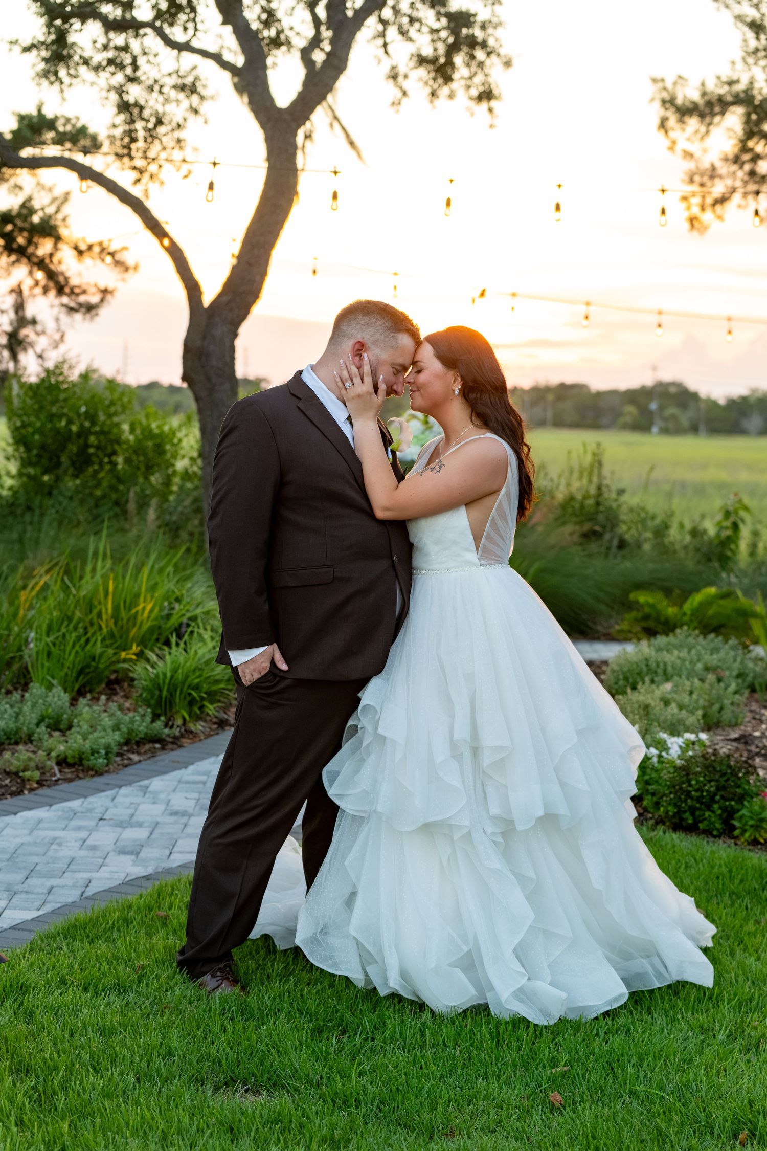 Bride and groom share a romantic sunset kiss in a lush garden, she wearing a layered white ball gown.