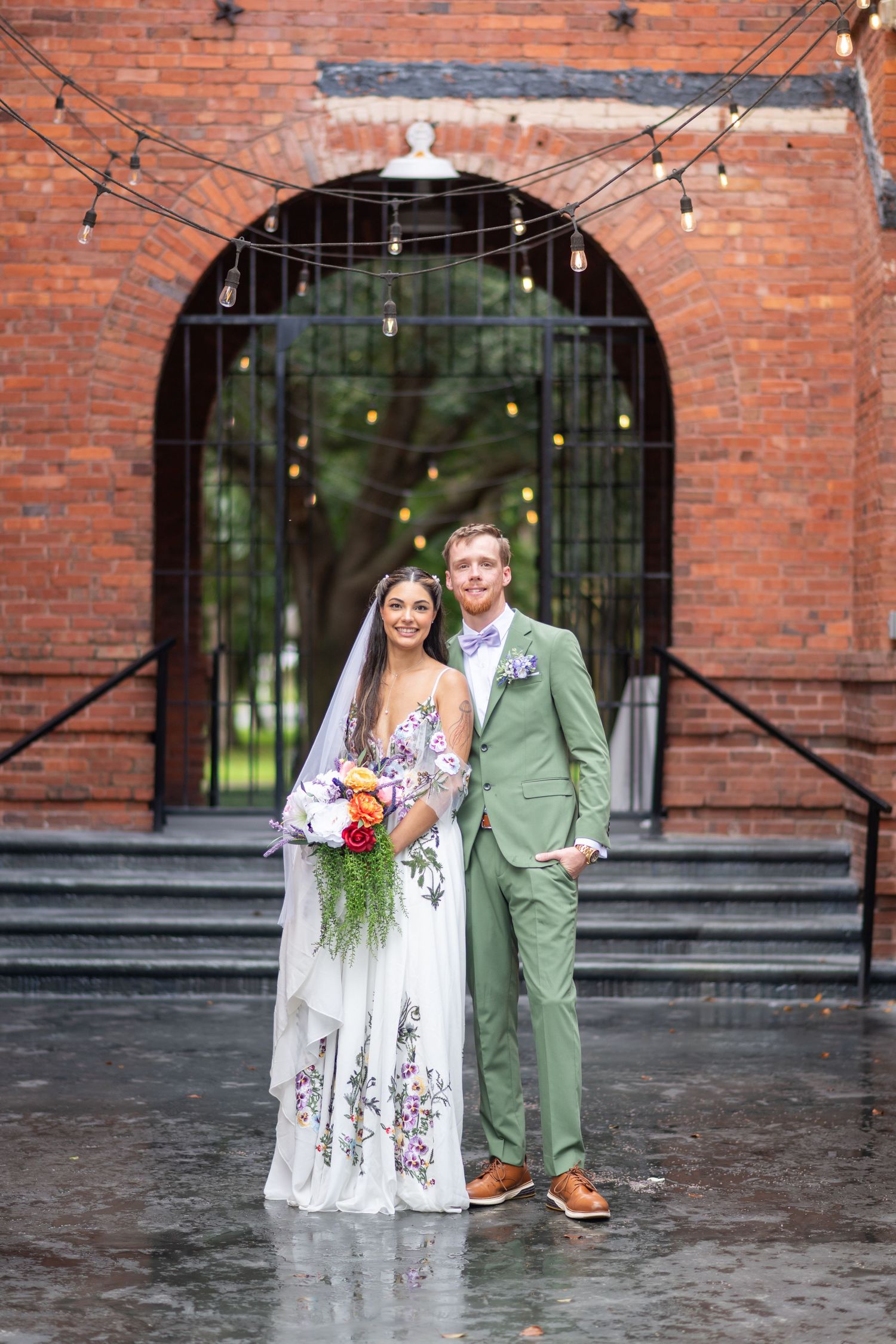 Smiling bride and groom posing before brick archway with string lights, she in floral gown, he in green suit.