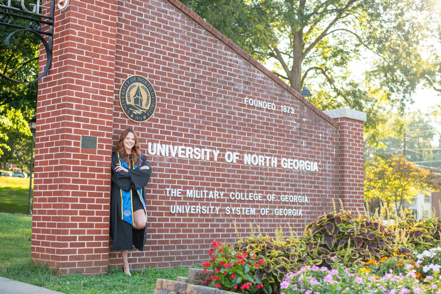 Graduate in cap and gown poses by University of North Georgia brick entrance sign surrounded by flowers.