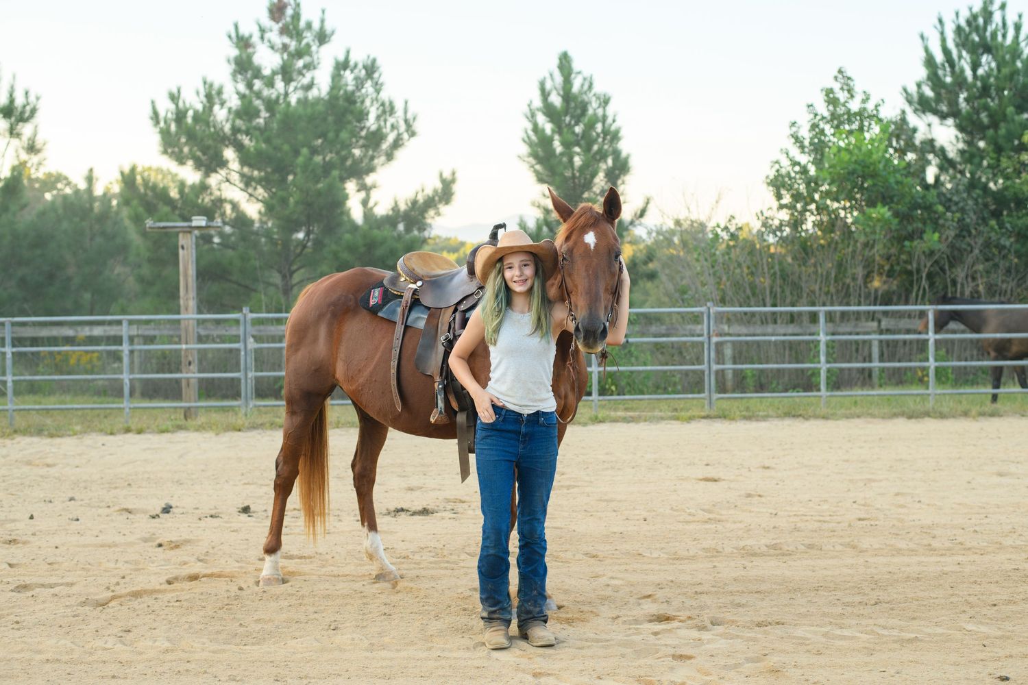 Young girl in cowboy hat standing beside a saddled chestnut horse in a sandy ranch arena with green trees.