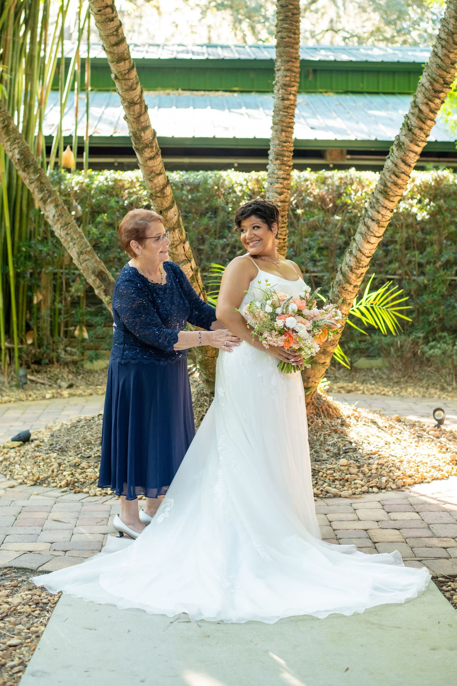 Bride in white gown holds floral bouquet beside woman in navy dress, standing among palm trees at outdoor wedding venue.