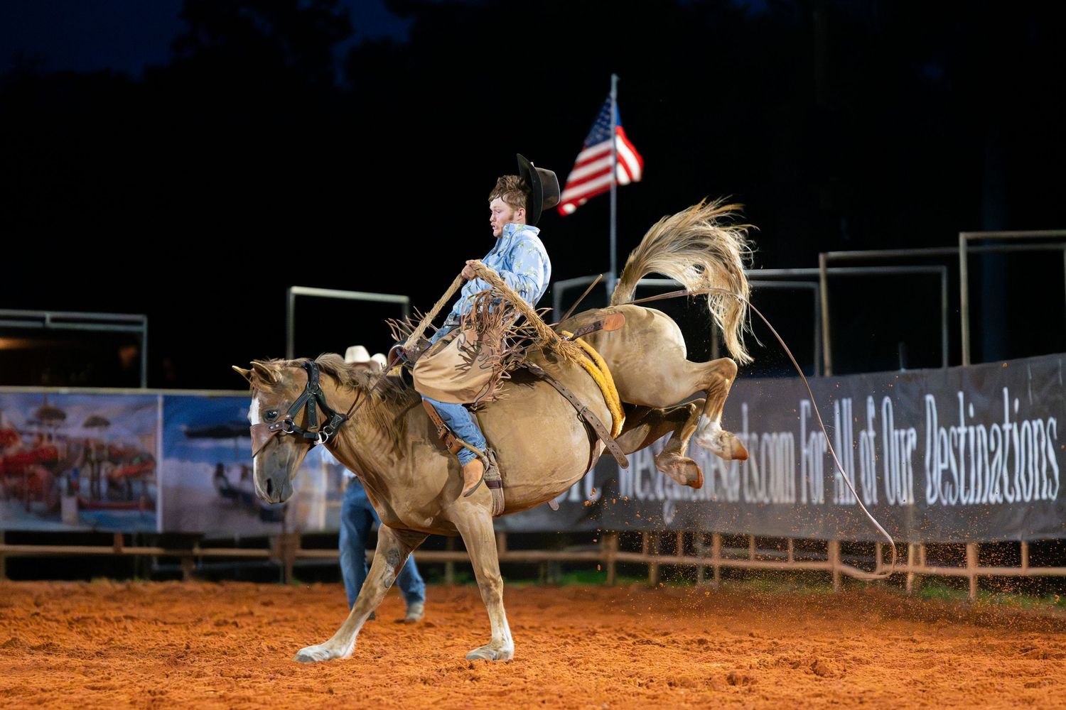 Cowboy riding a bucking bronco at a nighttime rodeo with an American flag in the background.