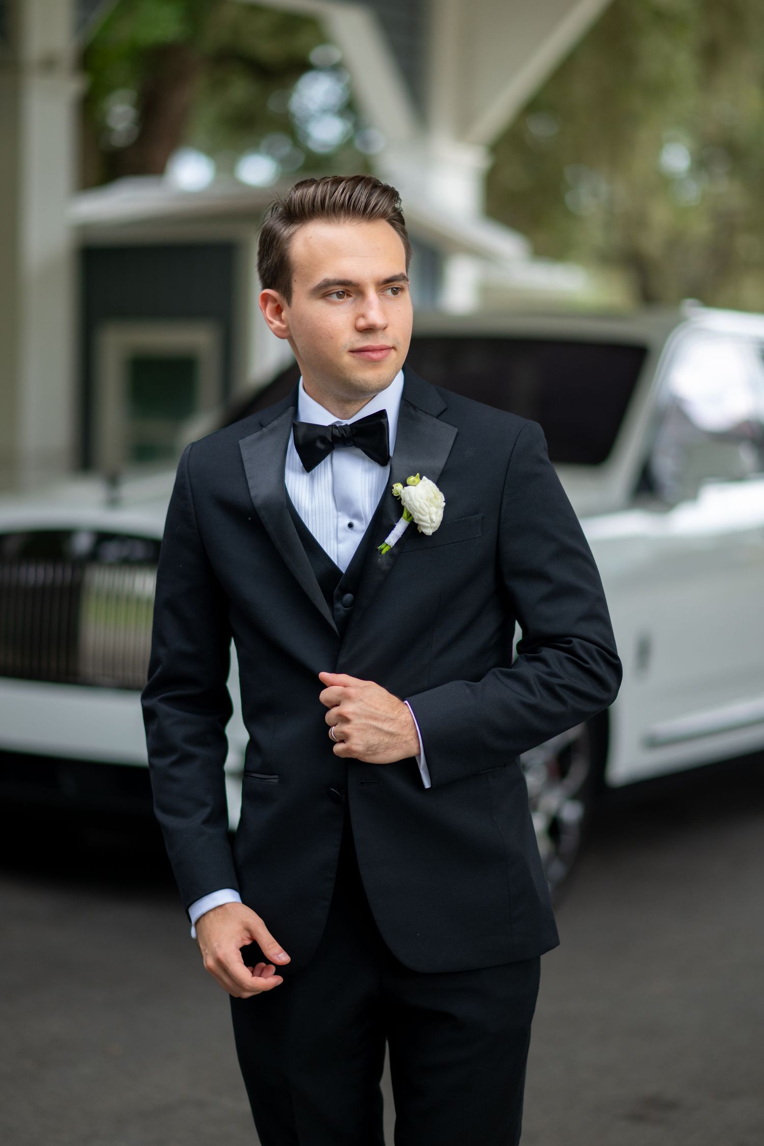 Young groom in black tuxedo with white boutonniere stands confidently outdoors near a white luxury car.