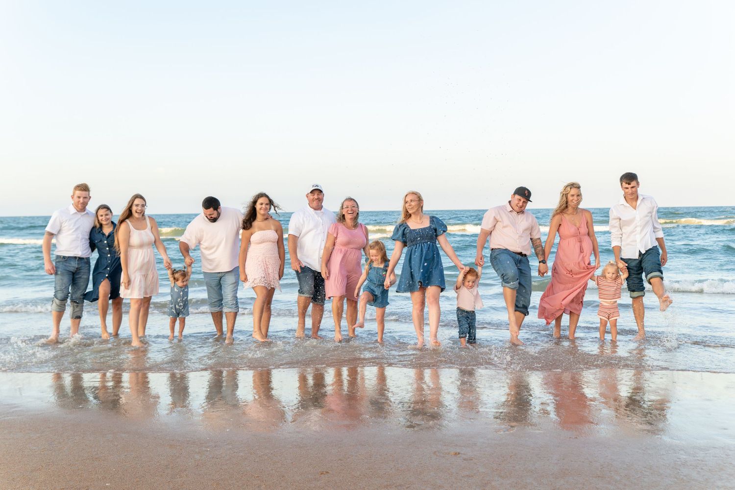Large family group holding hands on a sandy beach at sunset, wearing pastel outfits with ocean waves at their feet.