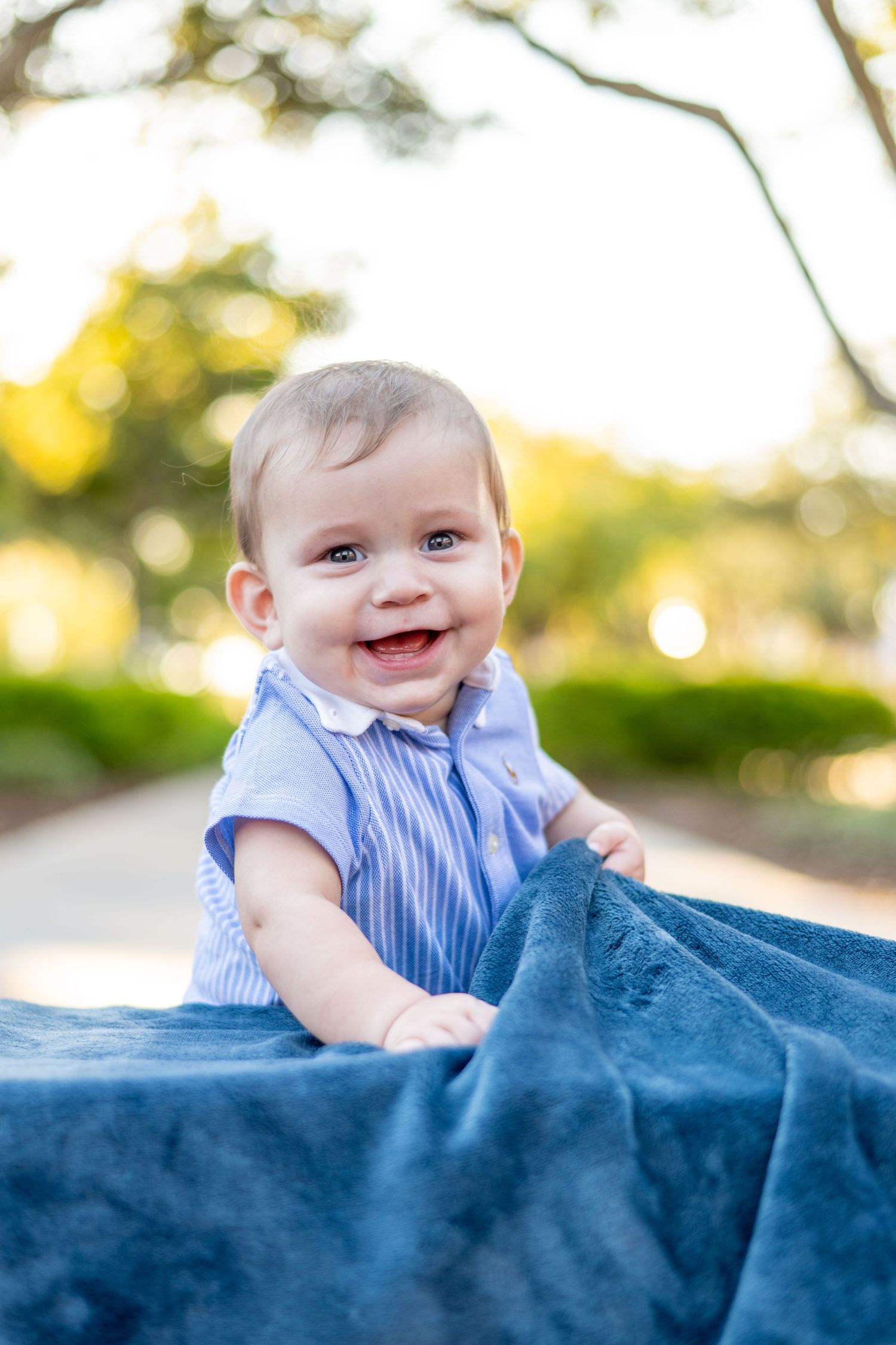 Adorable smiling baby in a blue outfit sitting on a soft blue blanket in a sunny park.