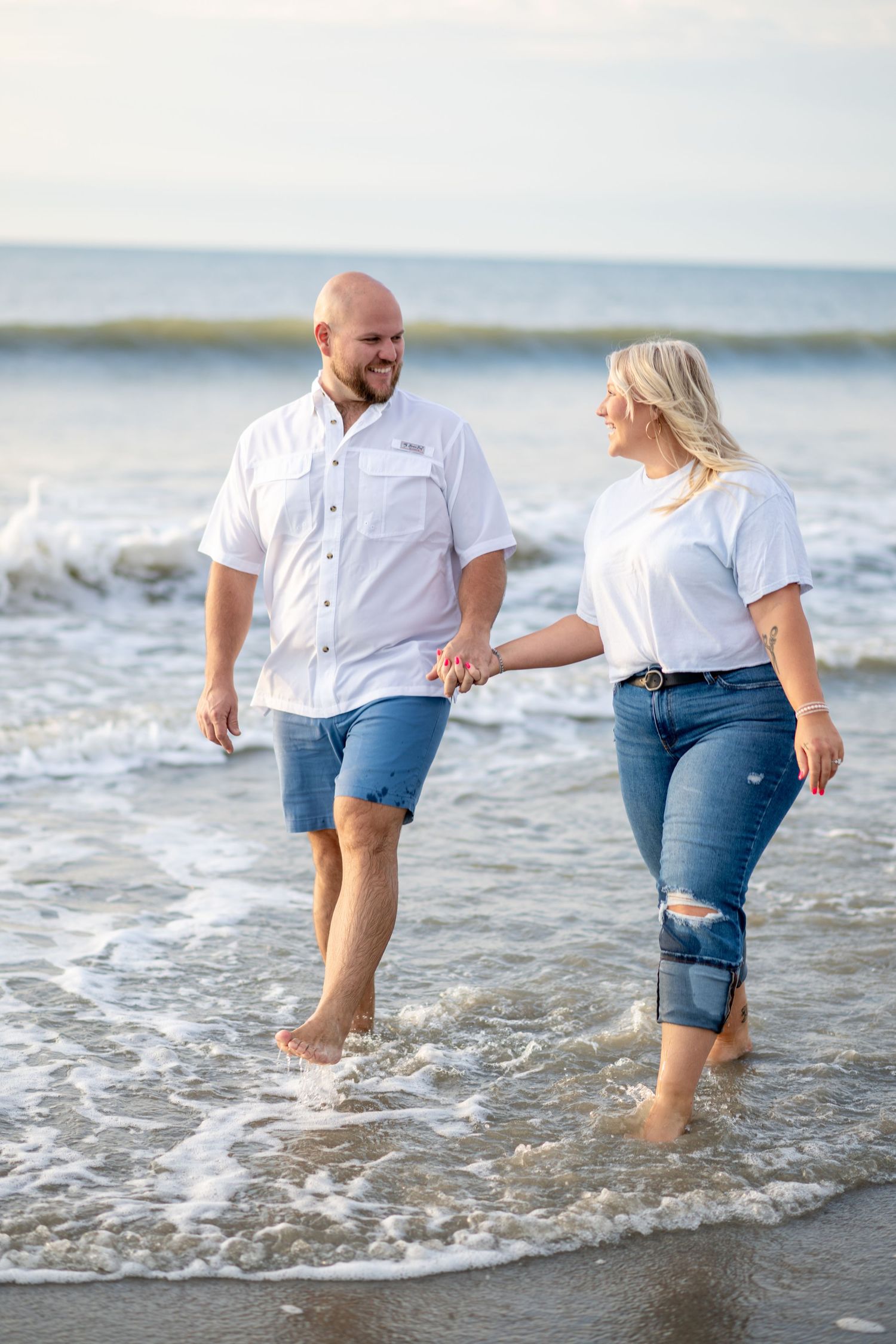 Happy couple holding hands and walking barefoot through ocean waves on a sunny beach day.