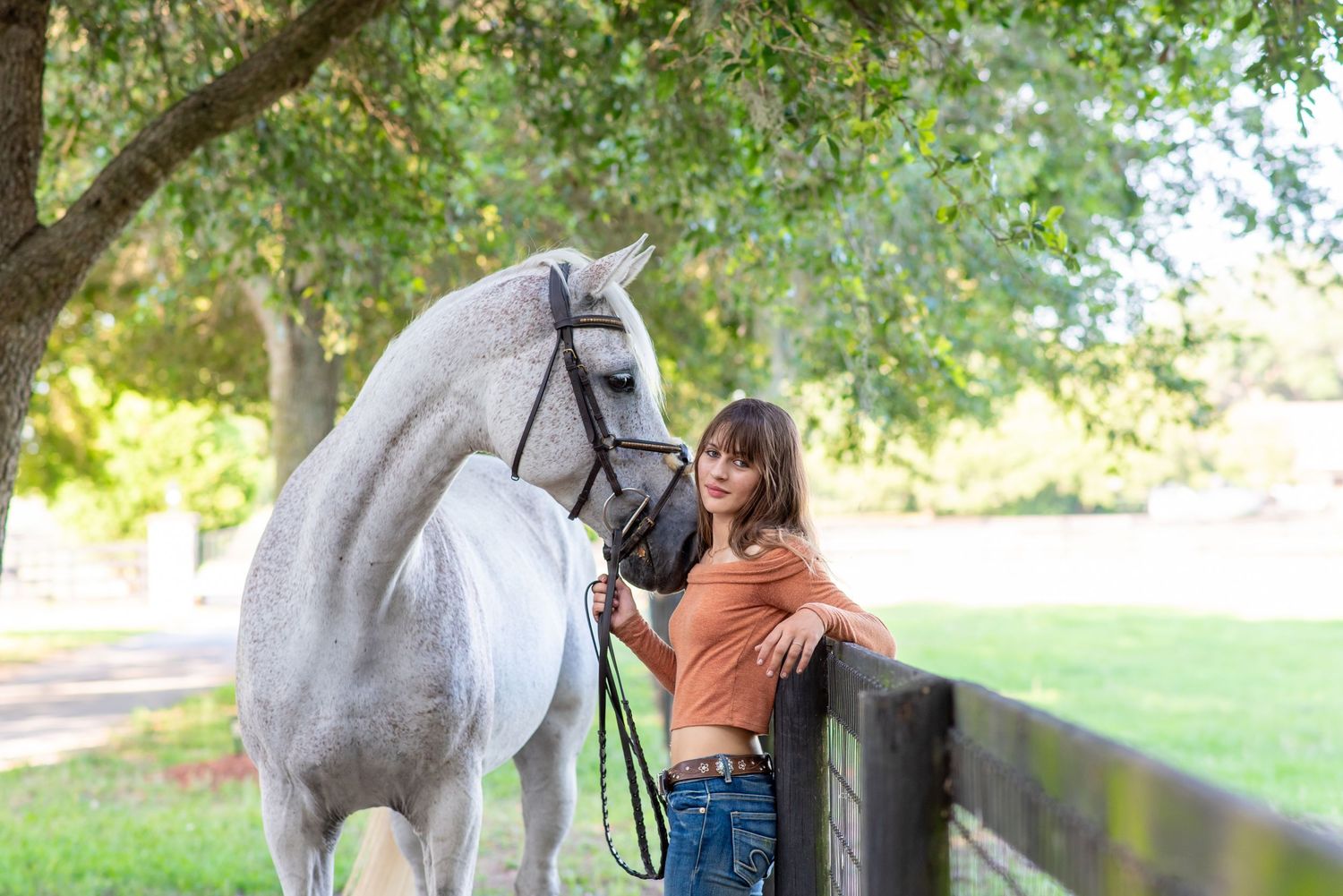 Young woman in orange top leaning on fence beside a white grey horse under trees.