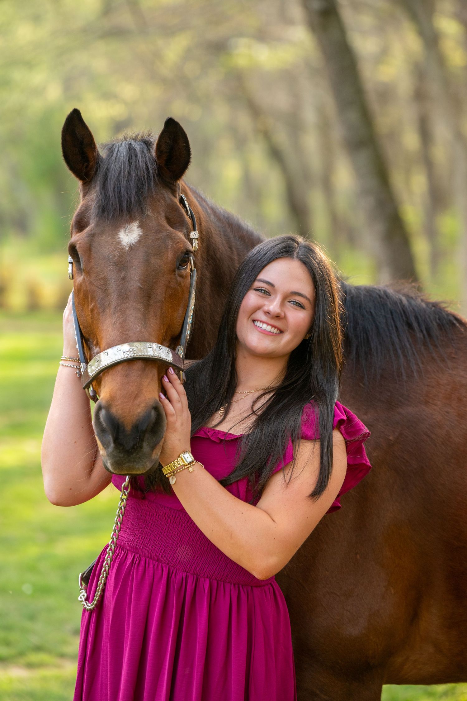 Smiling young woman in magenta dress posing with a bay horse outdoors in a lush green wooded setting.