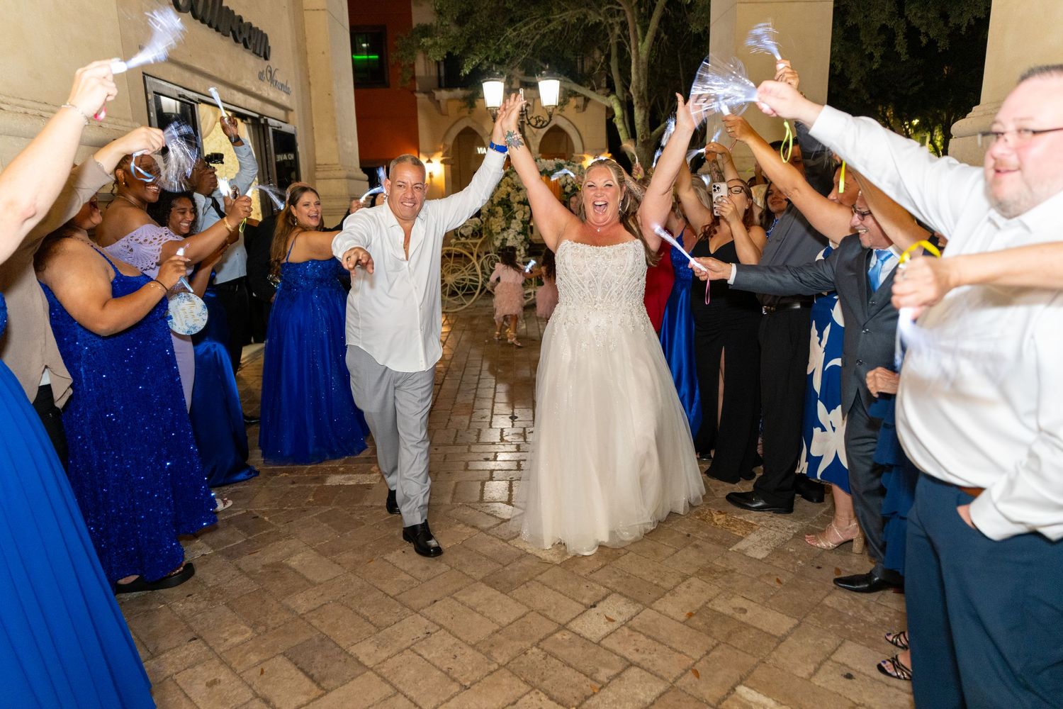 Bride and groom exit wedding reception through cheering guests in royal blue, raising hands in celebration.
