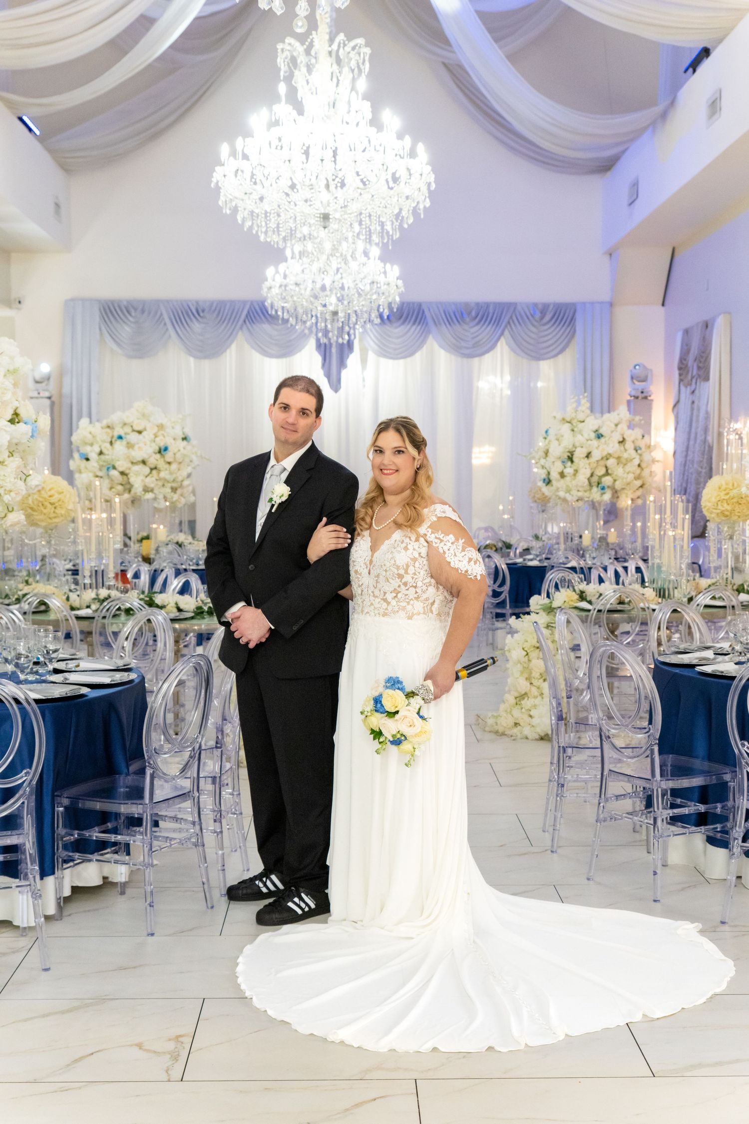 Bride and groom posing in elegant wedding reception hall with crystal chandelier, navy blue tables, and white floral centerpieces.