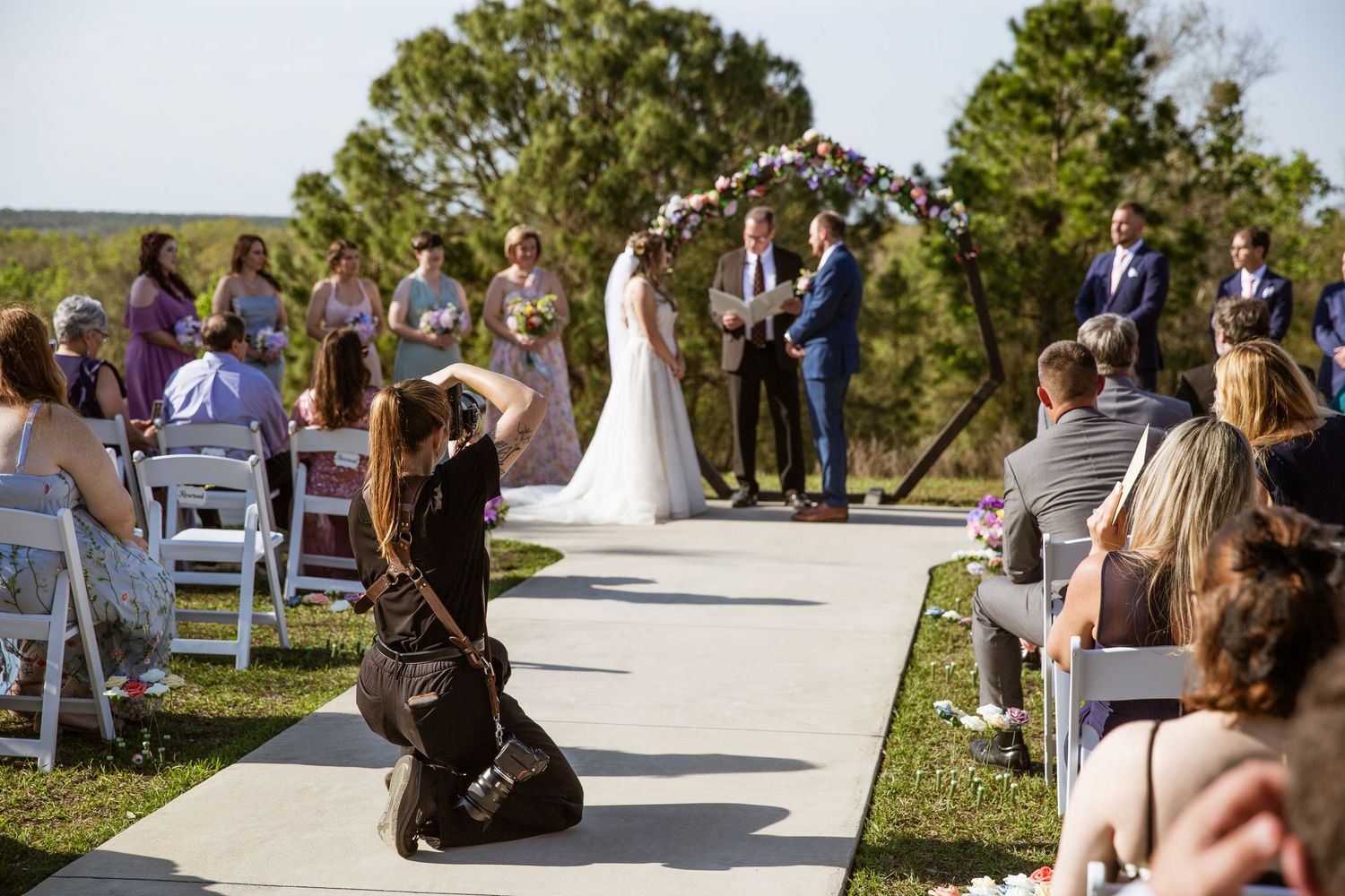Photographer kneels on aisle capturing bride and groom at outdoor wedding ceremony with floral arch.