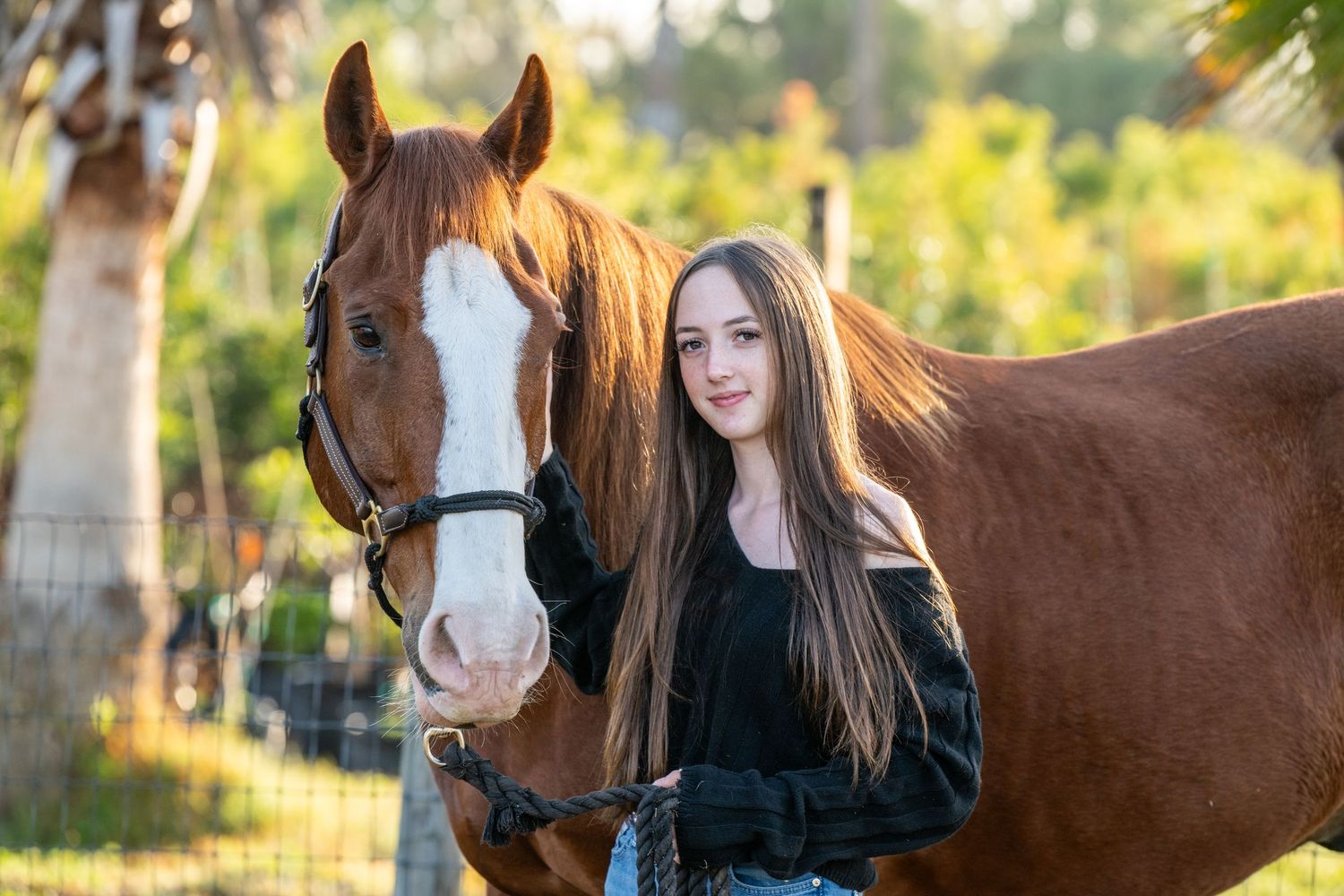 Young woman with long hair posing with a chestnut horse outdoors near a fence.