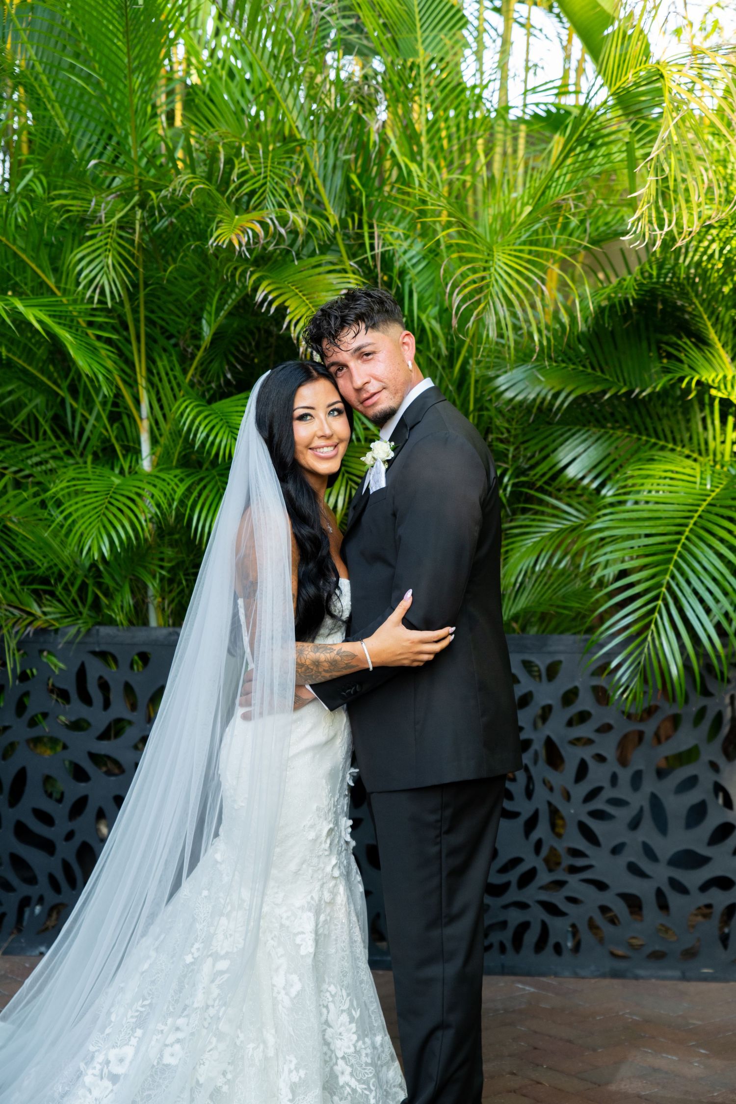 A bride and groom pose together in front of lush tropical palms, she wears a lace gown with veil, he in a black suit.