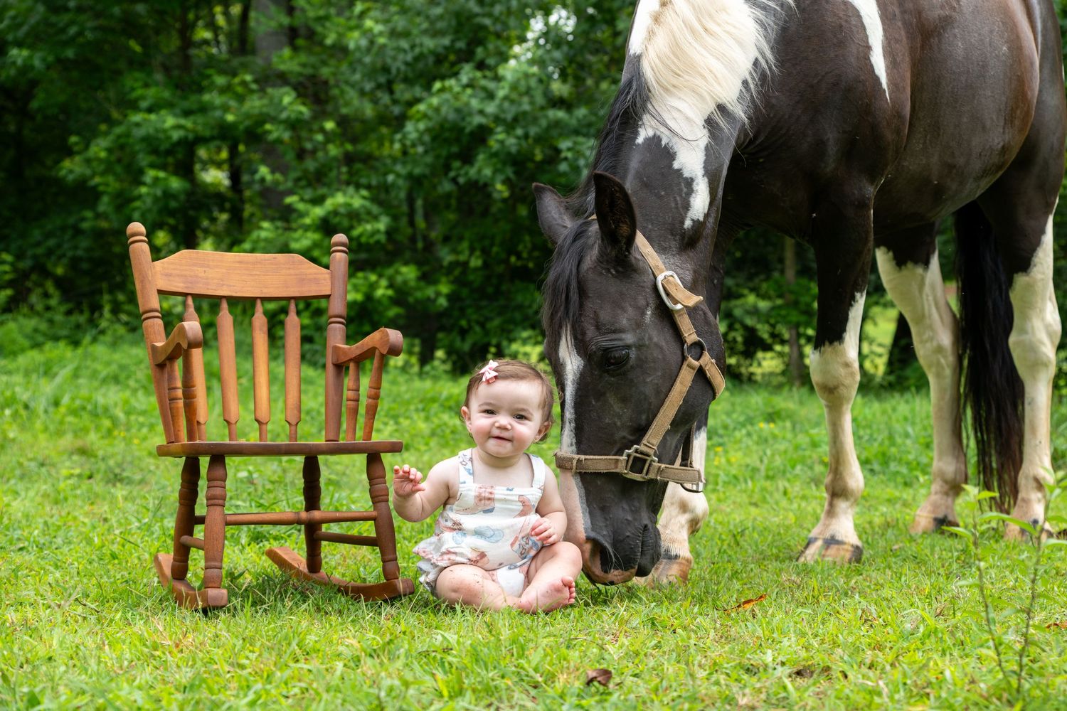 Baby girl sits on grass beside a wooden rocking chair and a grazing black and white horse outdoors.