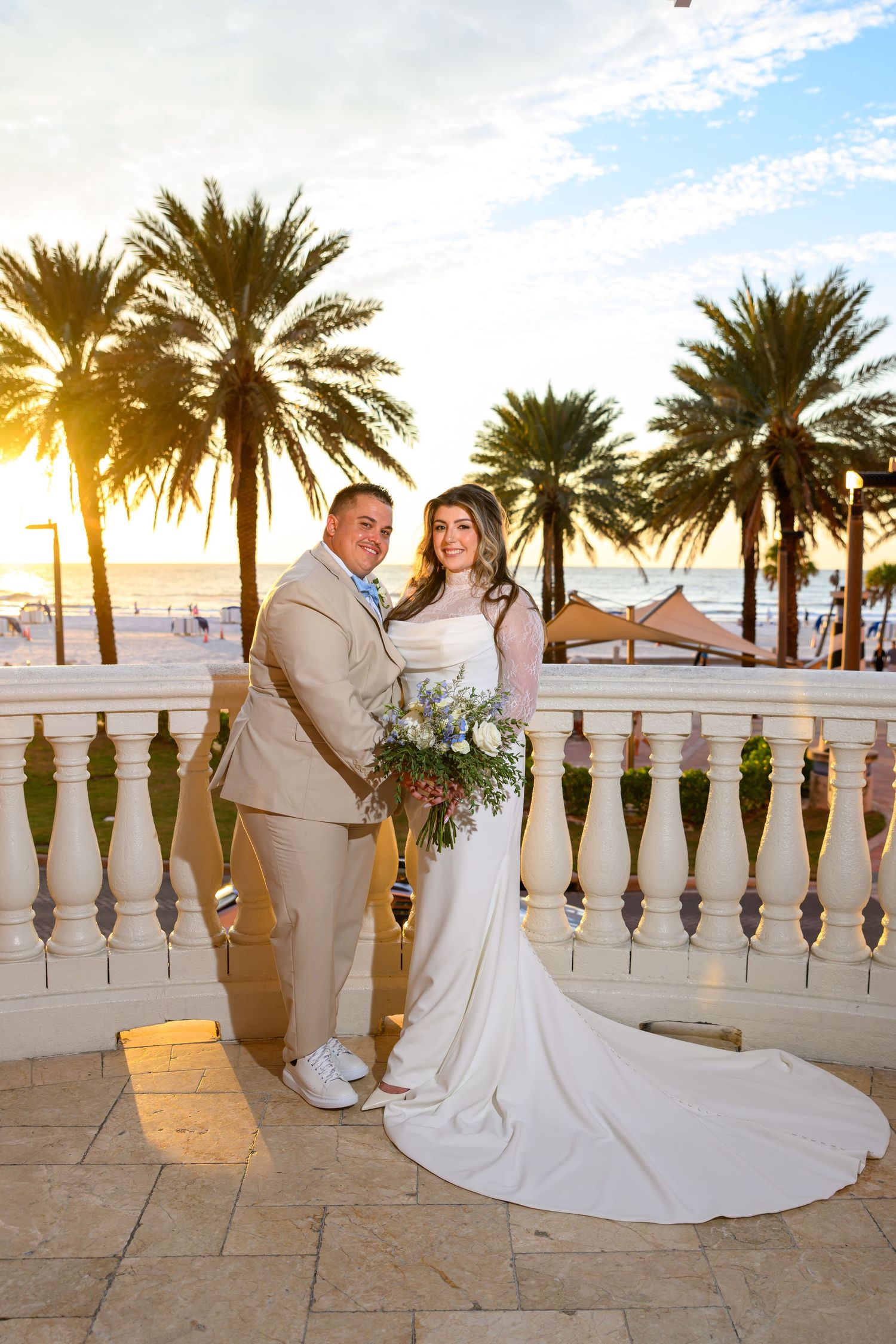 Bride in a white gown with a long train and groom in a tan suit pose with a bouquet on a beachfront terrace with palm trees at sunset.