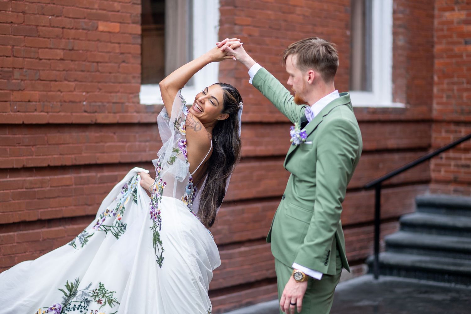 Bride and groom dancing outdoors near brick wall, she wears floral gown, he wears sage green suit.