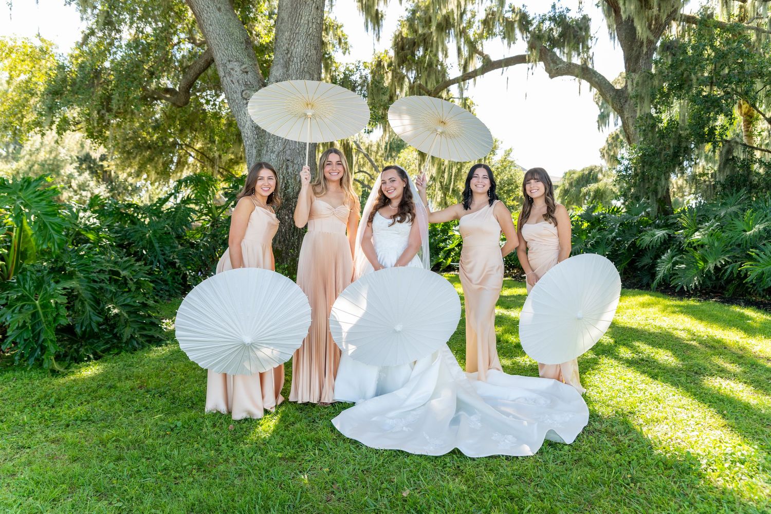 Bride and bridesmaids in champagne gowns posing with white parasols under a large oak tree.
