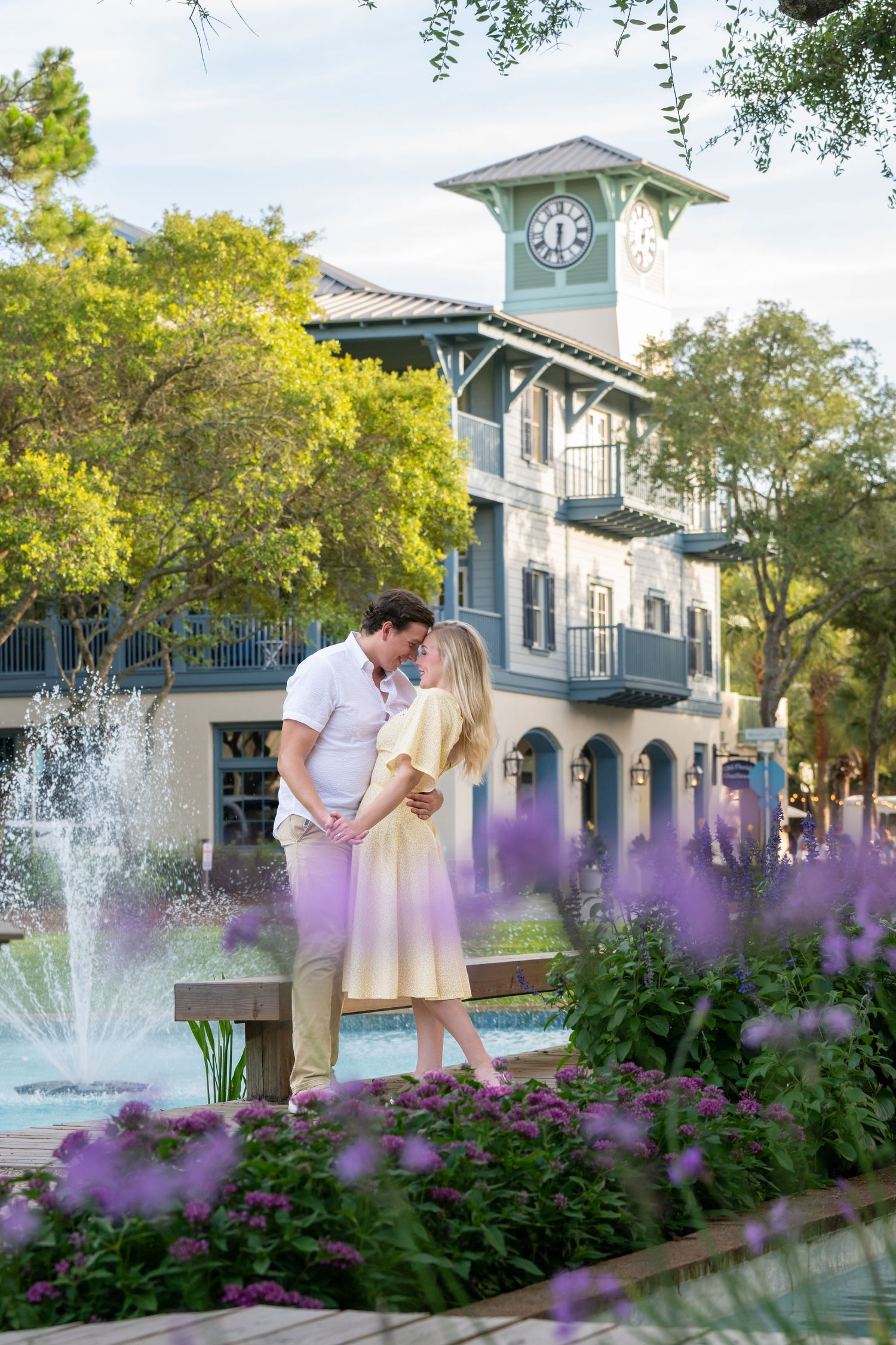 Couple sharing a romantic kiss near a fountain with purple flowers and a charming clock tower building in the background.