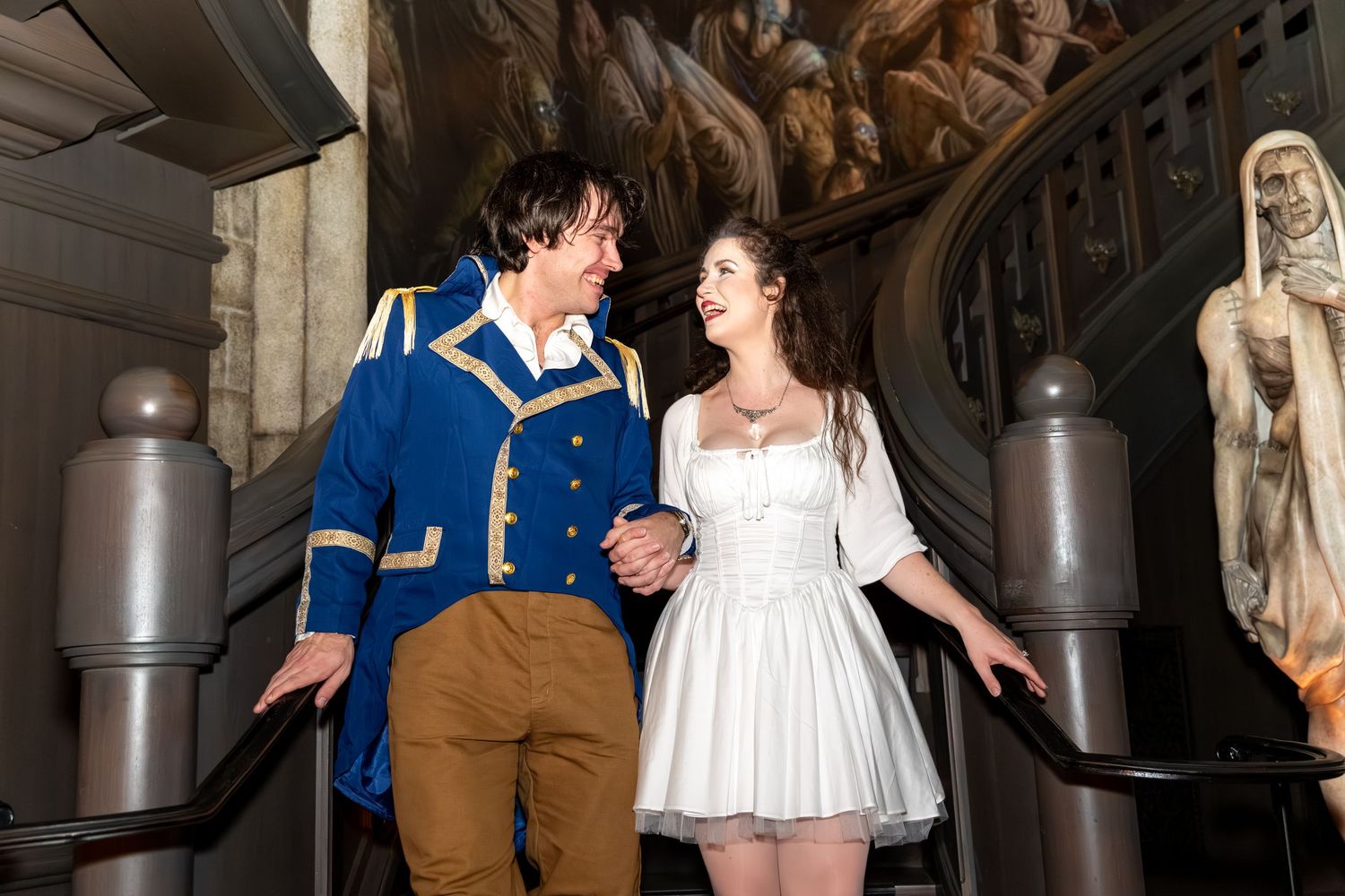 Couple in Beauty and the Beast costumes smiling on ornate staircase, woman in white dress, man in blue jacket.