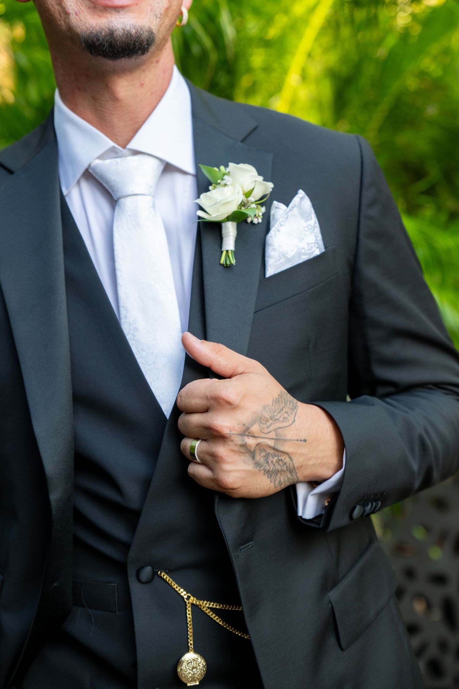 Groom in dark suit with white boutonniere, pocket square, gold pocket watch, and tattooed hand adjusting lapel.
